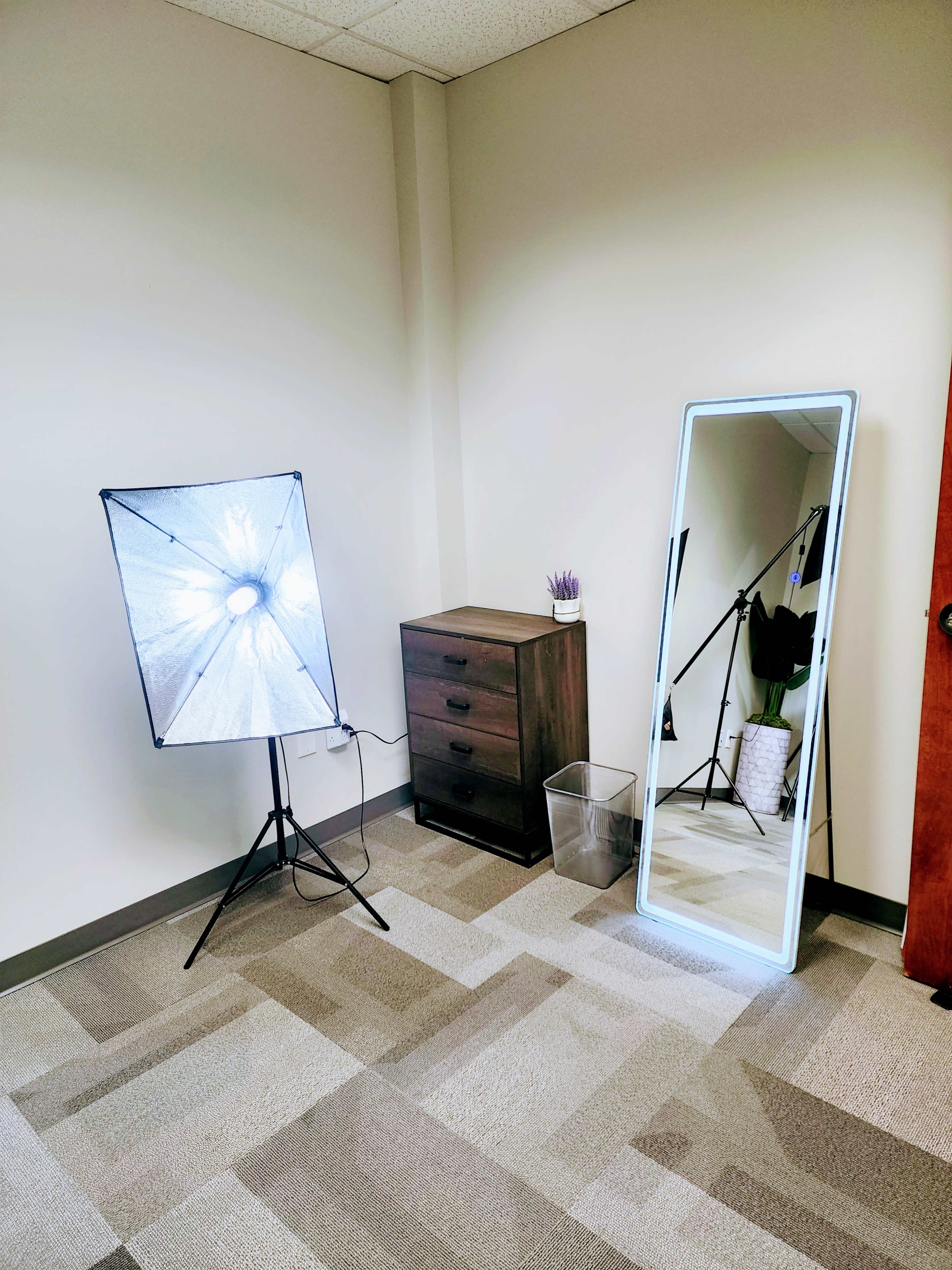 A corner of a room featuring a softbox light, a tall mirror, a small dresser with drawers, and a wastebasket.
