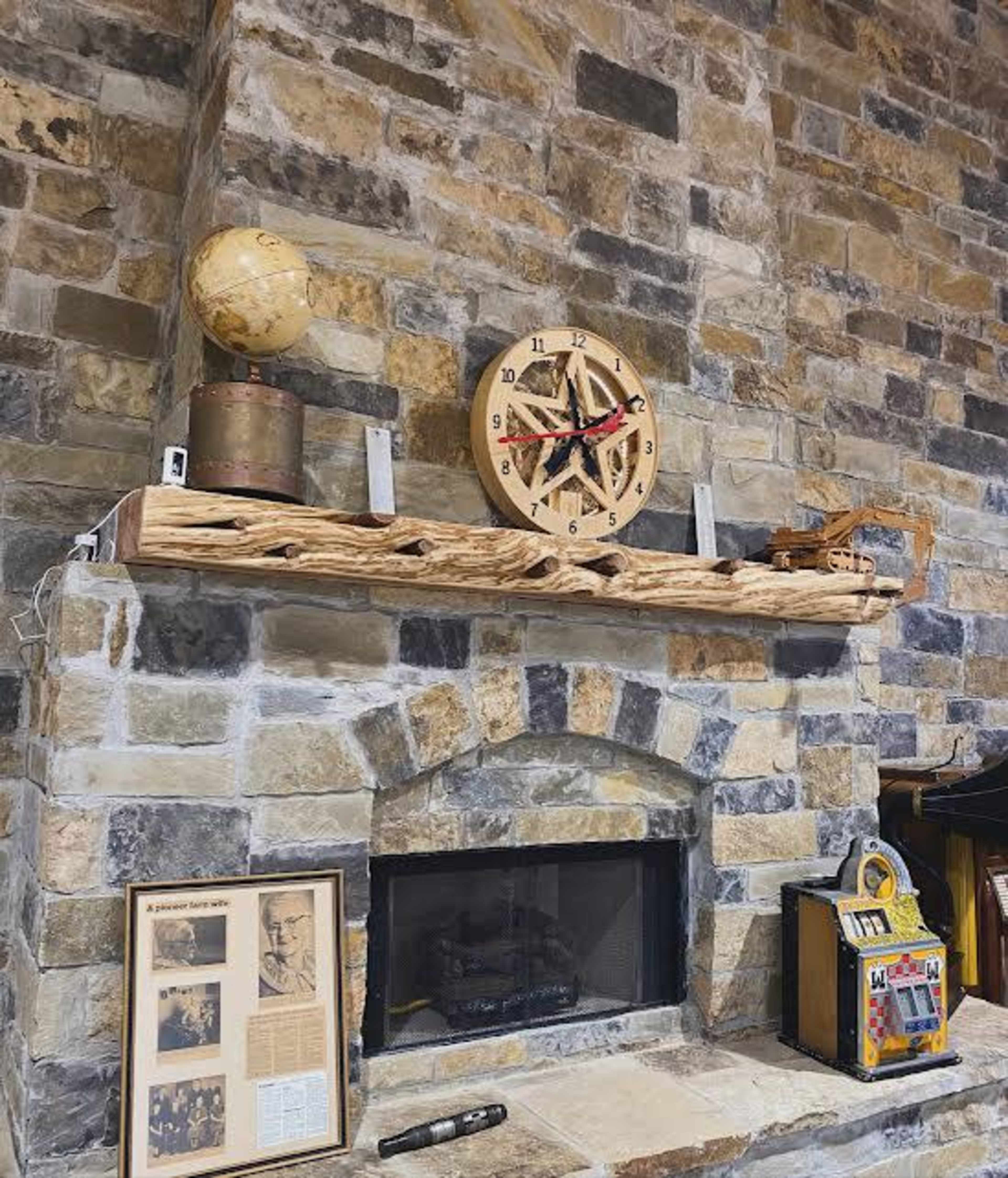 The image shows a rustic stone fireplace with a wooden mantel displaying a globe, a clock, and a small wooden ship model, alongside a framed photograph and a vintage-style jukebox.
