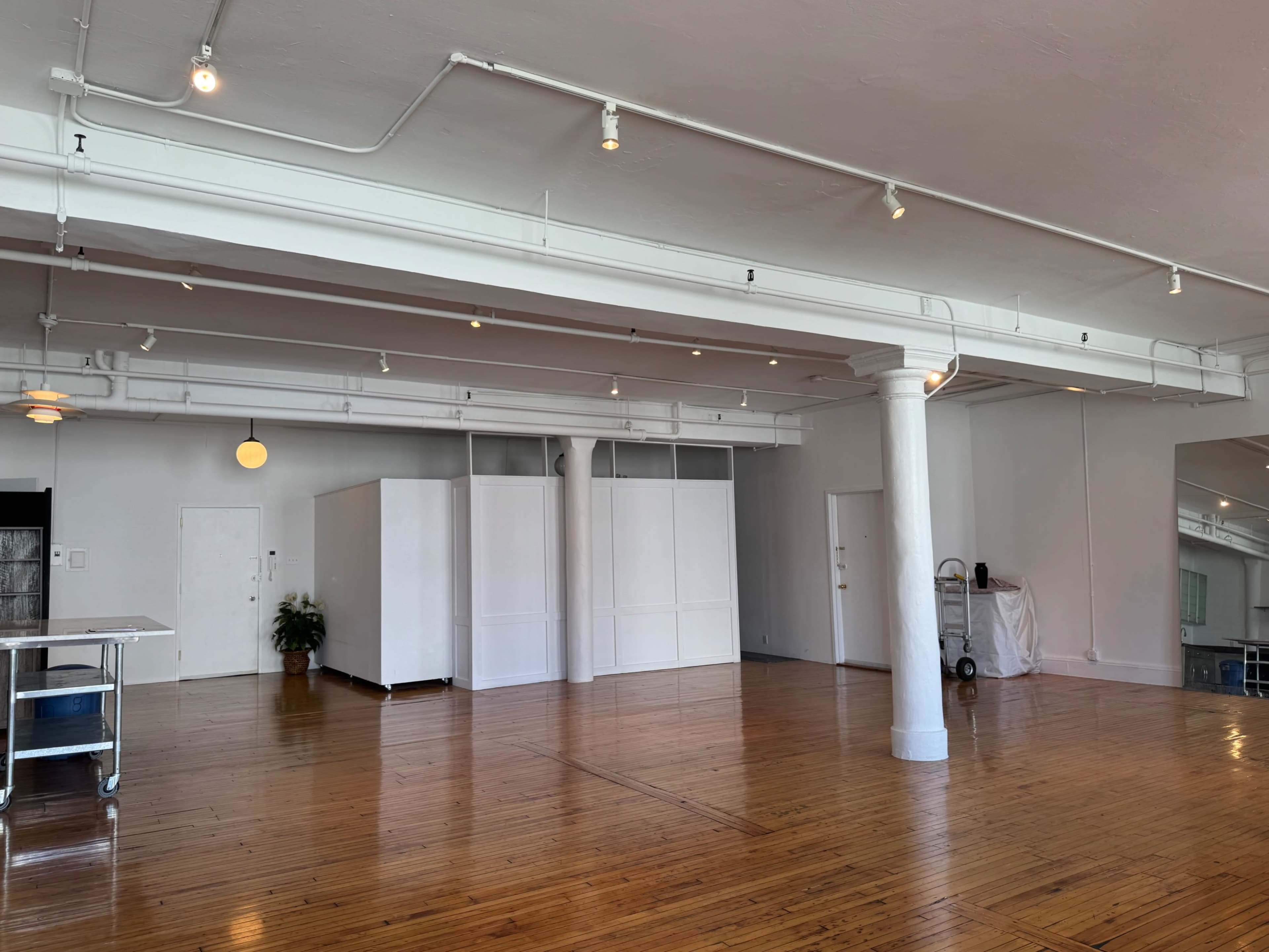 The image shows a spacious, empty room with wooden flooring, white walls, and overhead lighting.