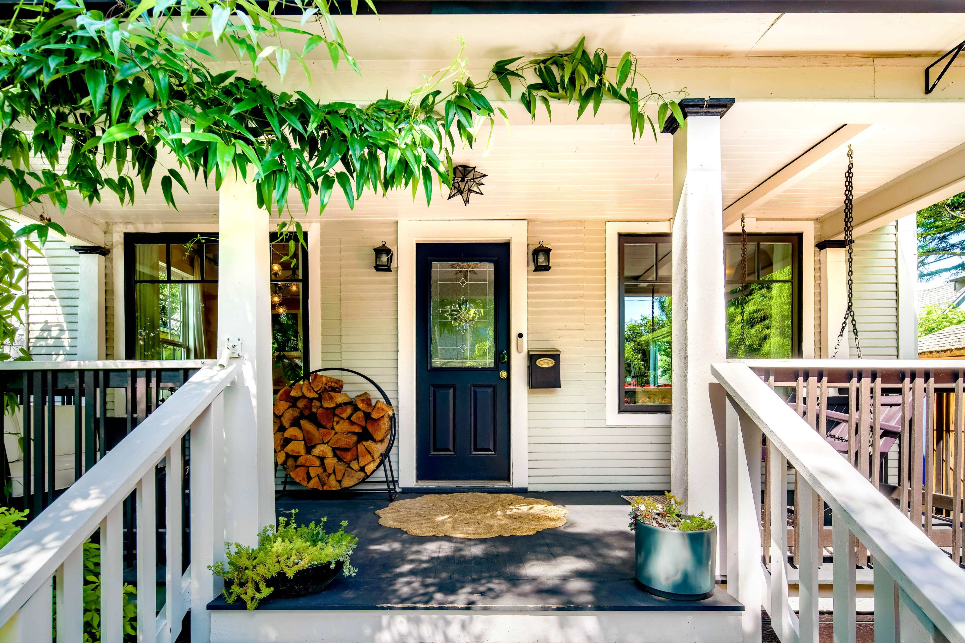 The image shows a front porch with a dark door, potted plants, a stack of firewood, and a welcome mat.
