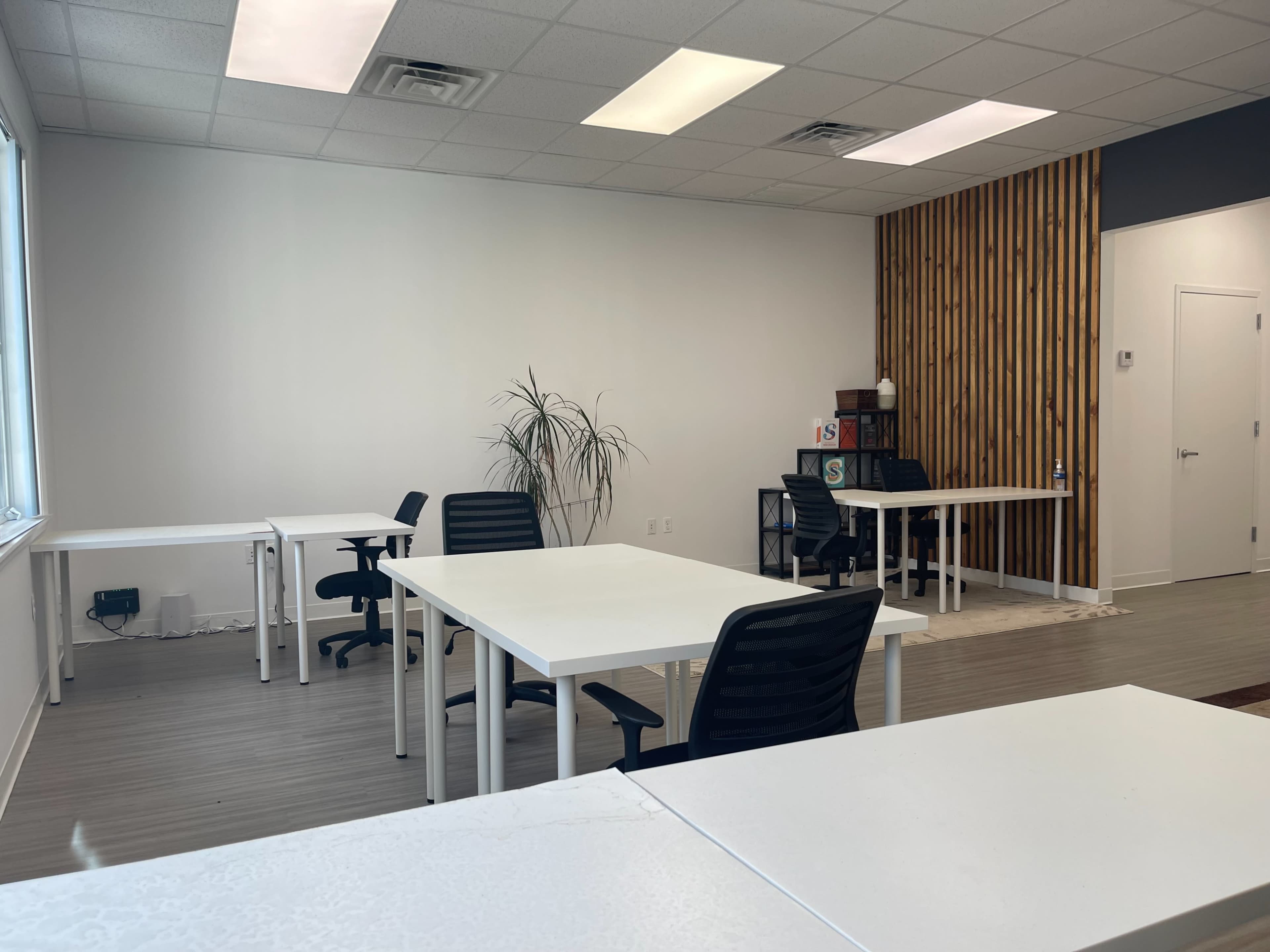 The image shows a spacious office with white tables and black chairs, a large window, and a wooden accent wall.