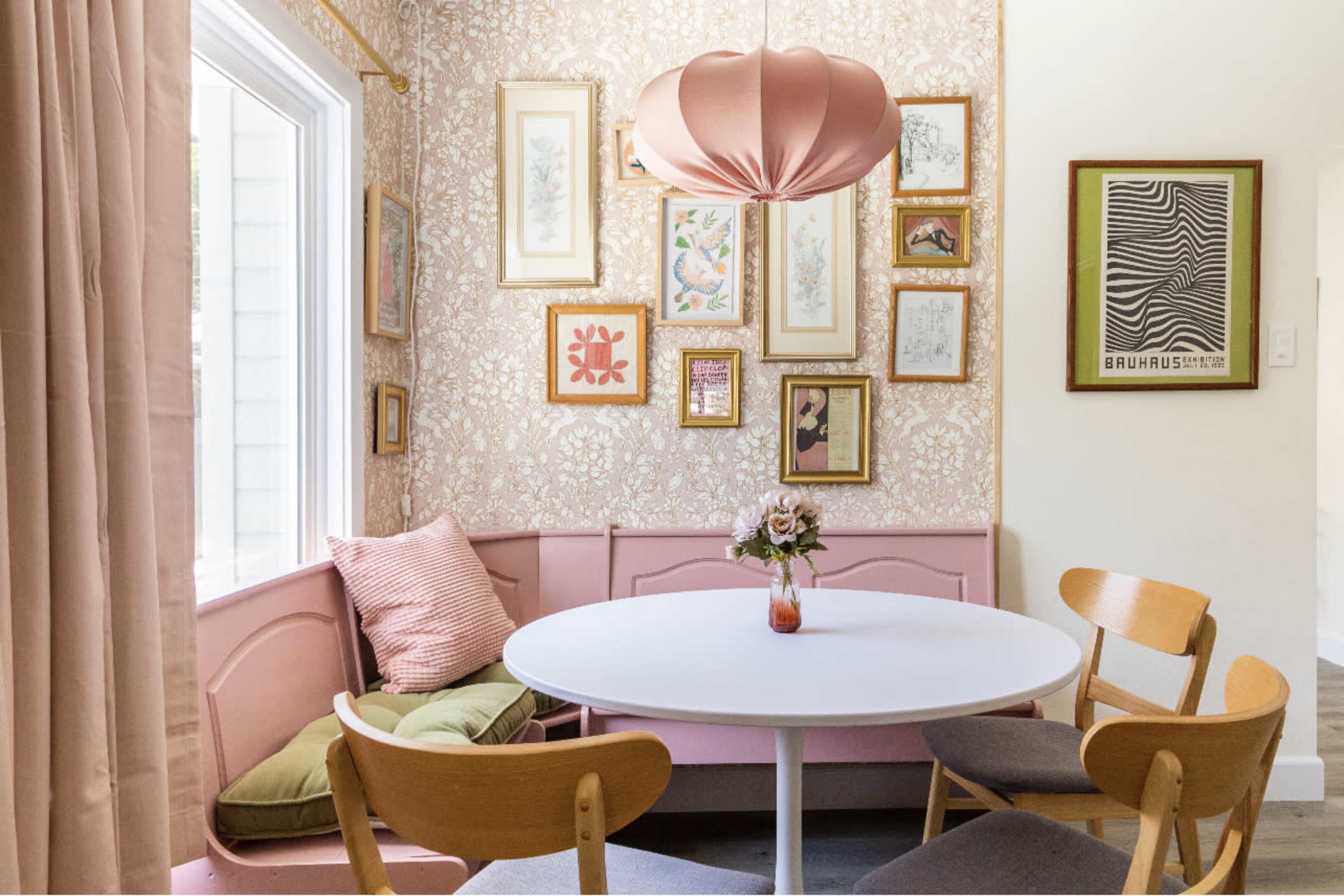 A round white table is set in a corner nook adorned with pink walls, framed artwork, and a pendant light.