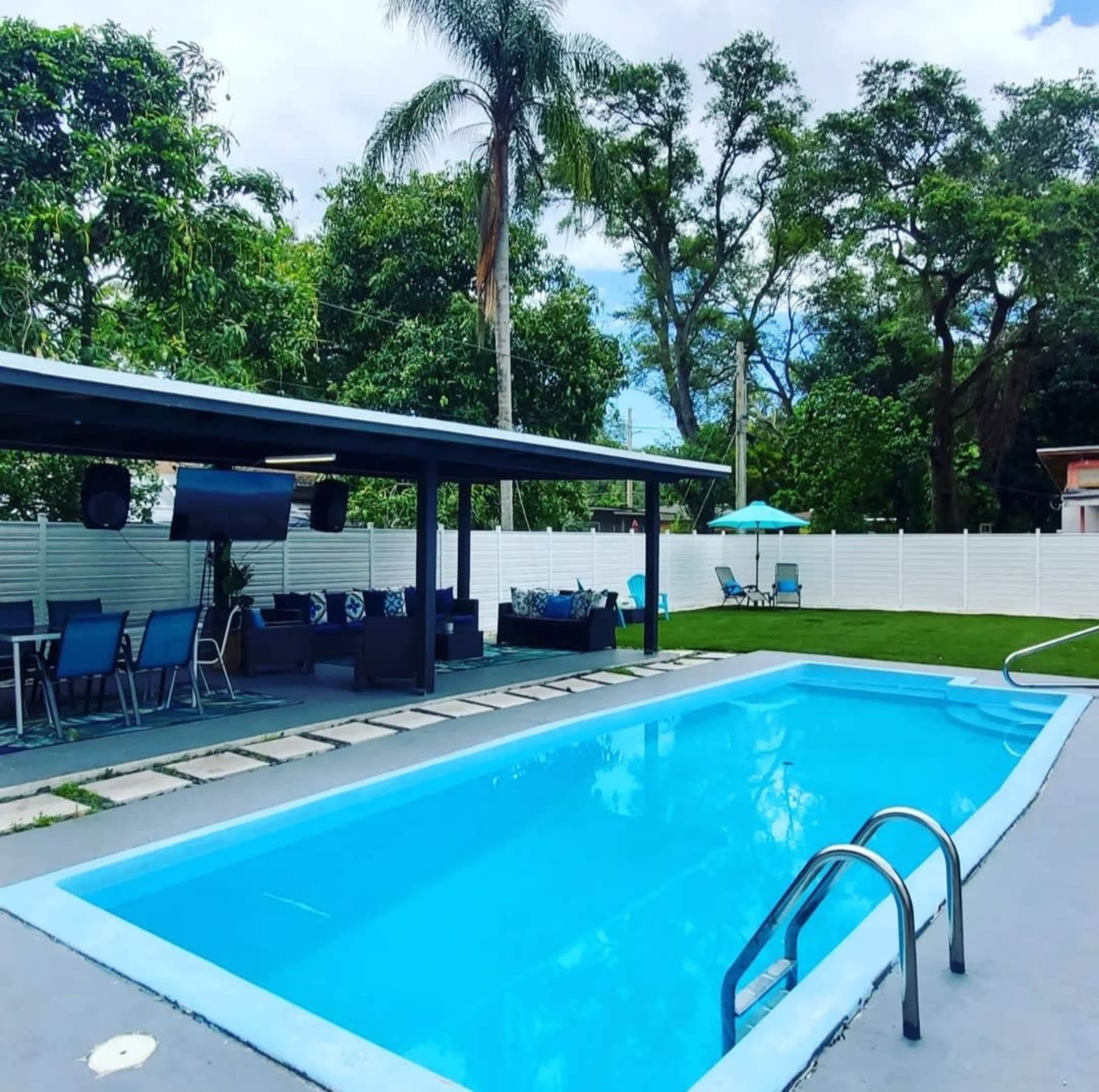 A swimming pool surrounded by trees and patio furniture, with a covered seating area nearby.