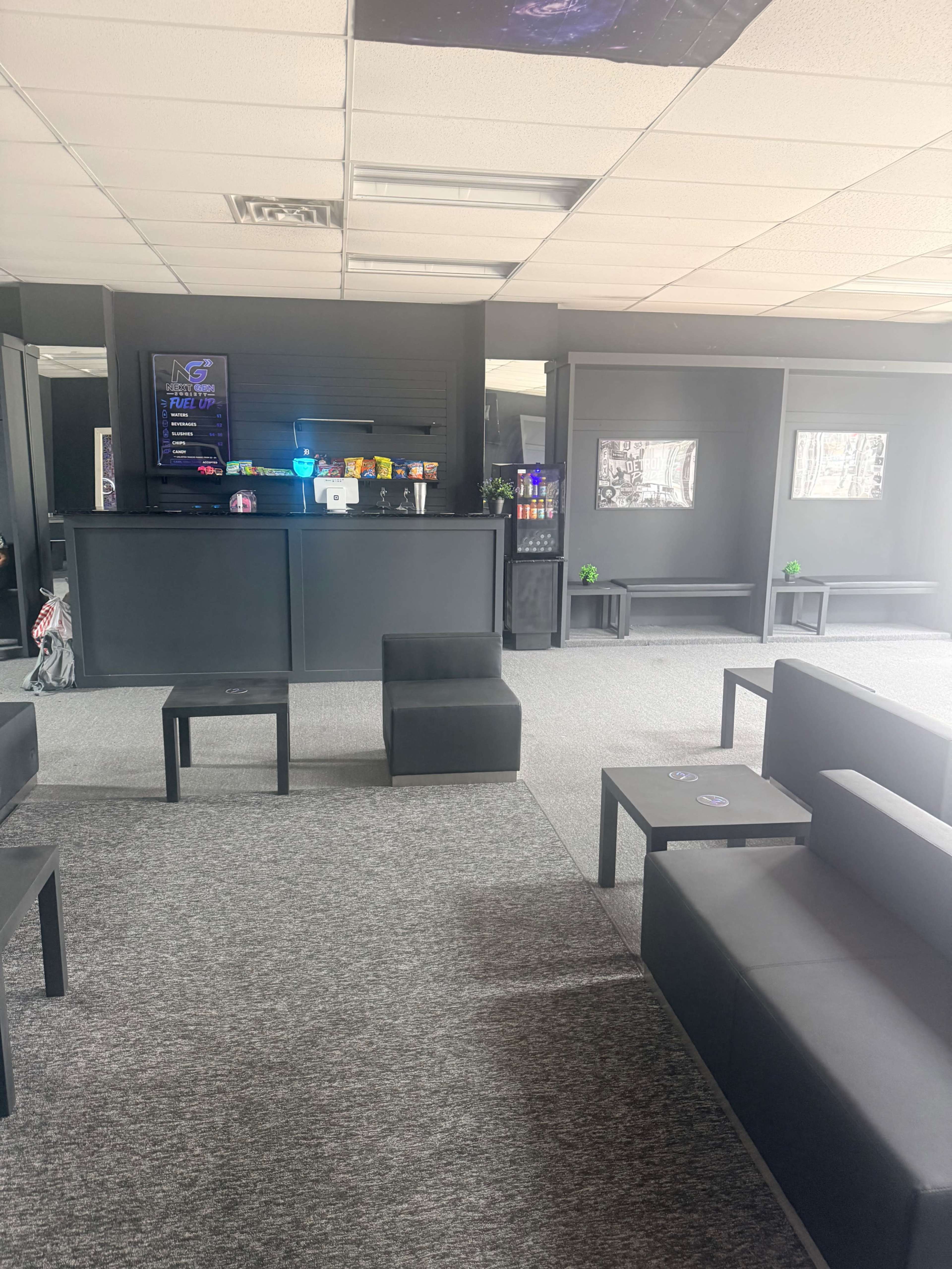 The image shows a modern lounge area with black furniture, including seating and tables, and a counter displaying colorful items in a well-lit space.
