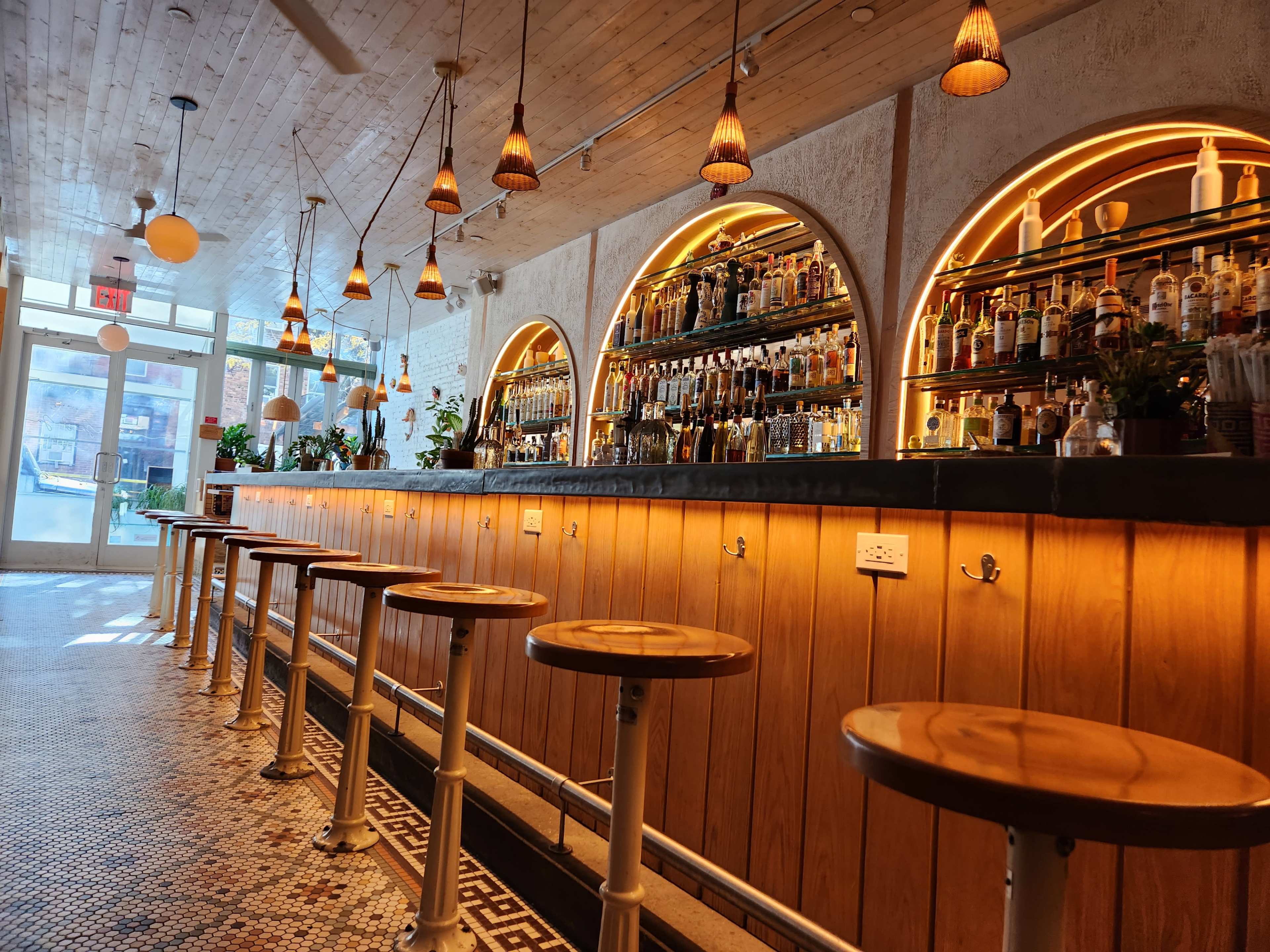 A bar with wooden stools and a well-stocked bar shelf illuminated by warm lighting, set in a spacious, well-lit room.