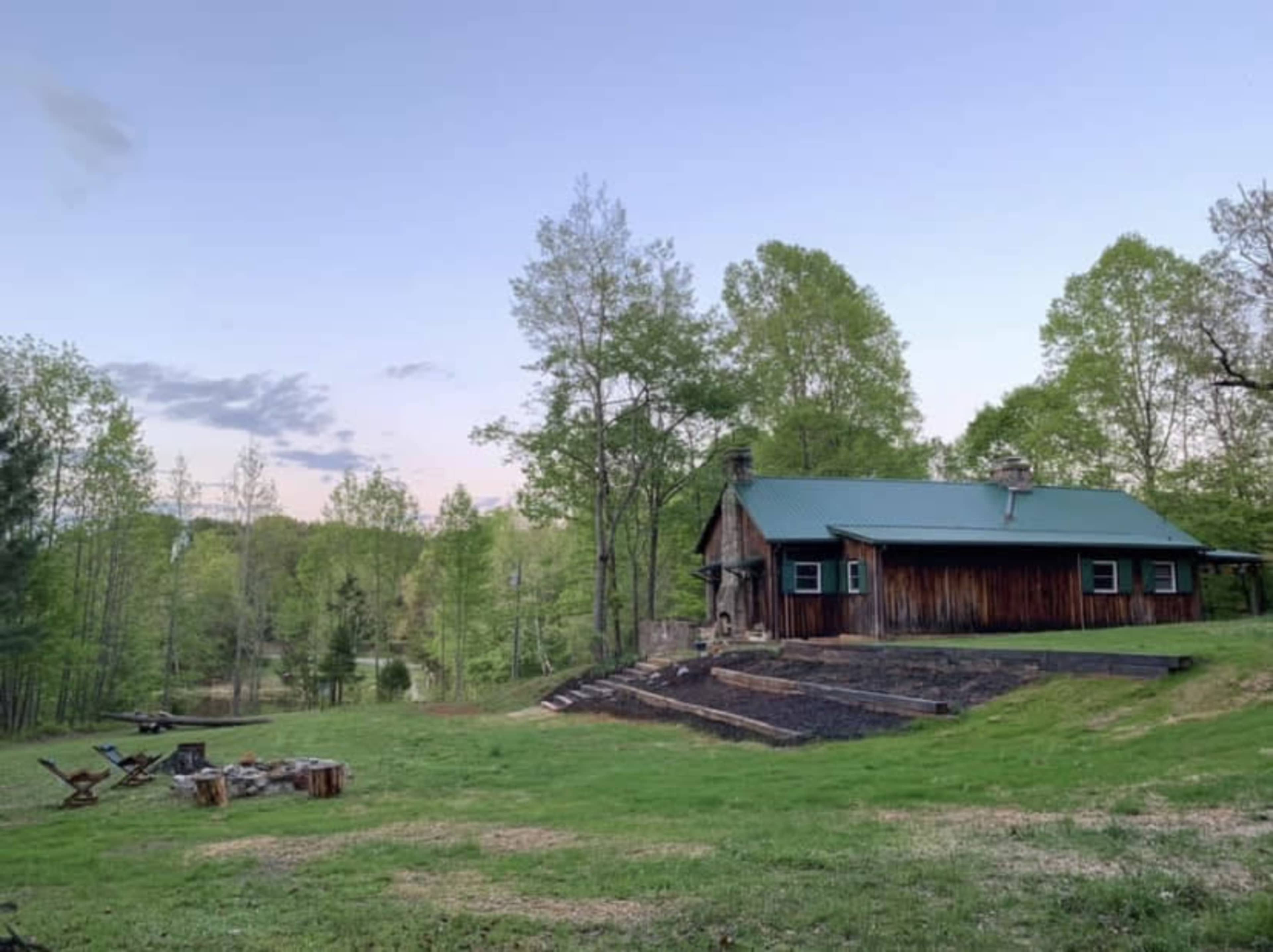 A wooden cabin with a green metal roof sits on a grassy hillside surrounded by trees and a fire pit area.