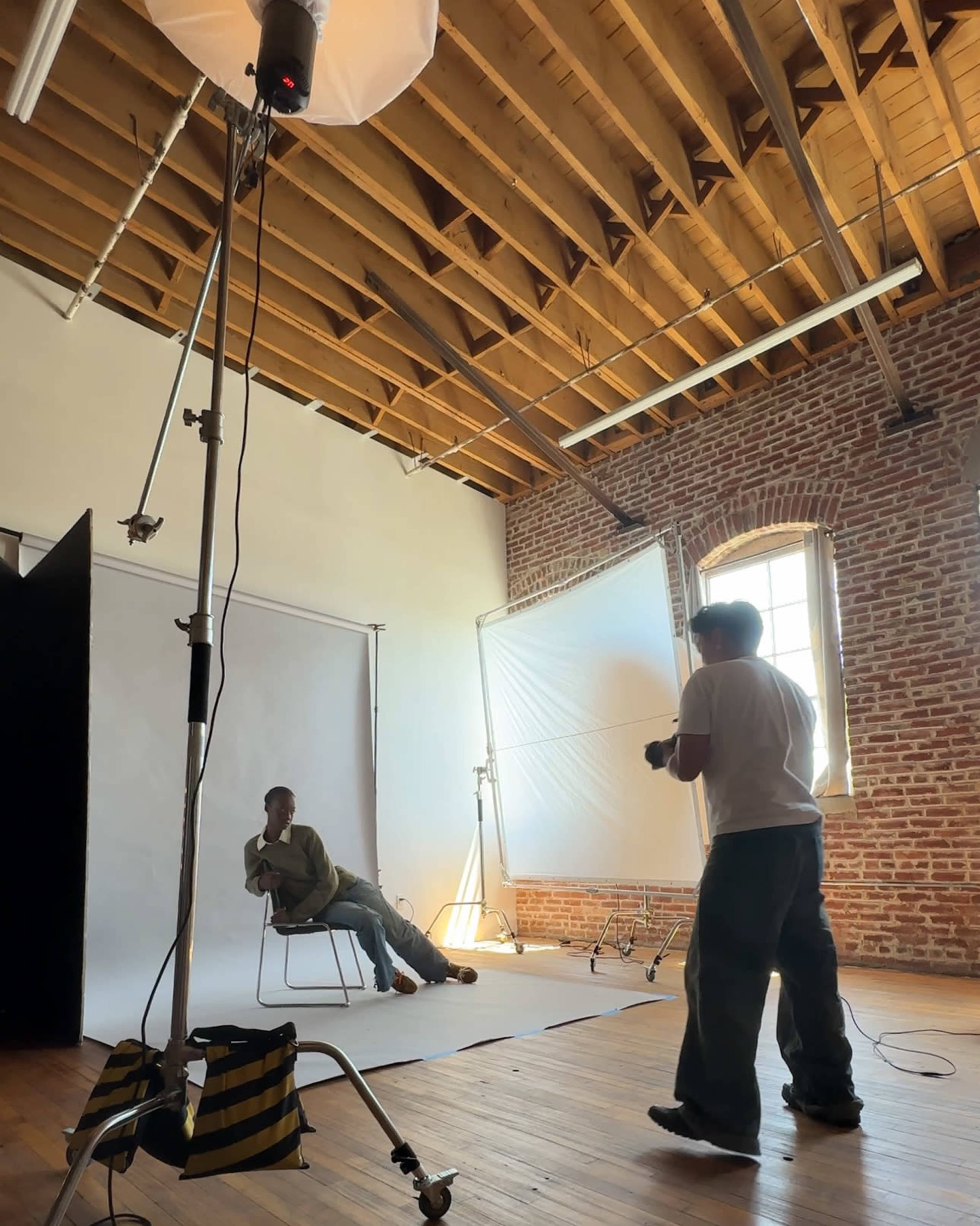 A person is sitting on a chair in a well-lit studio while another individual is preparing to take a photograph.