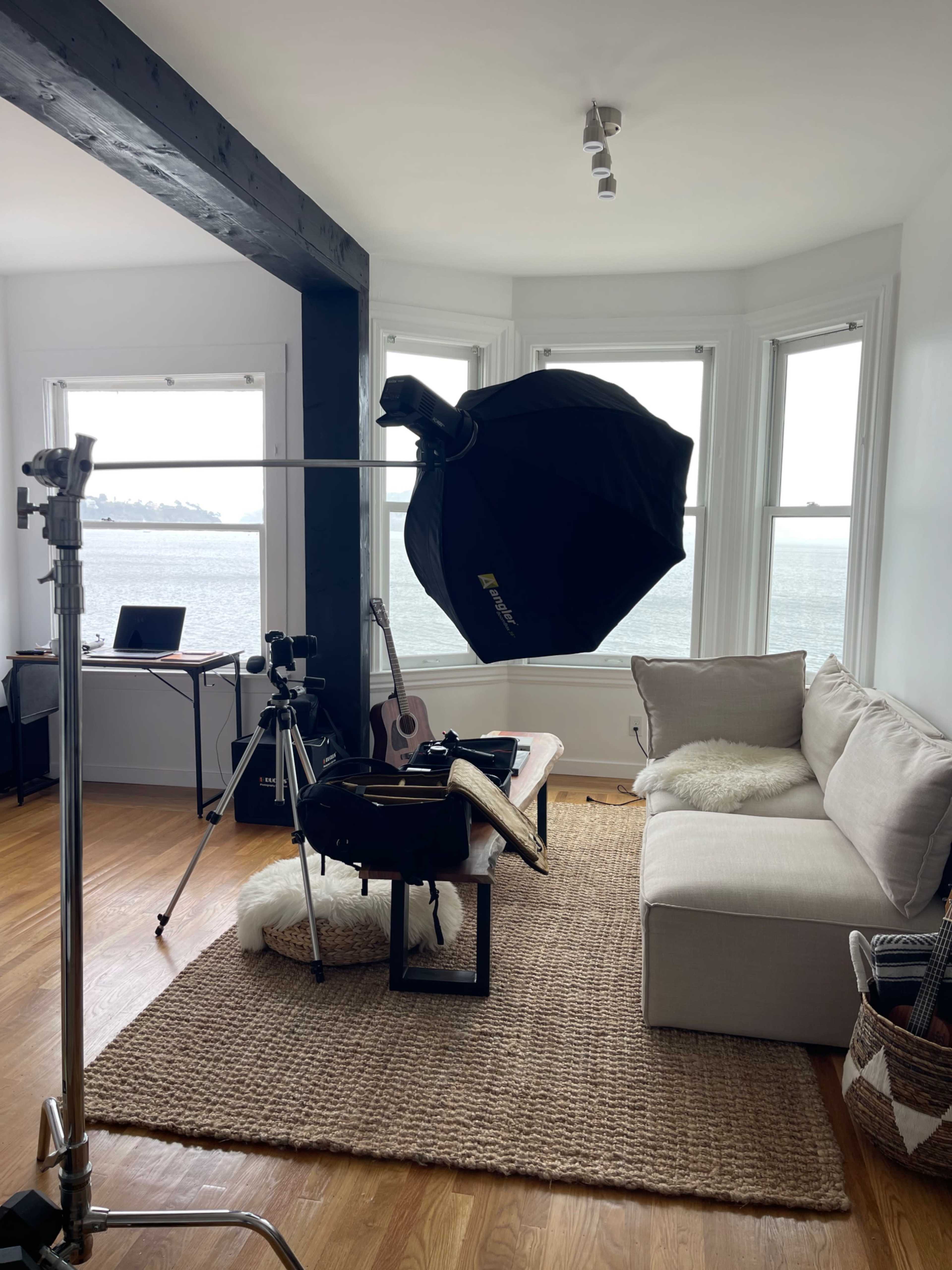 A photography setup is arranged in a sunny room with panoramic windows, featuring a softbox lighting equipment, a couch, and various instruments and equipment on a table.