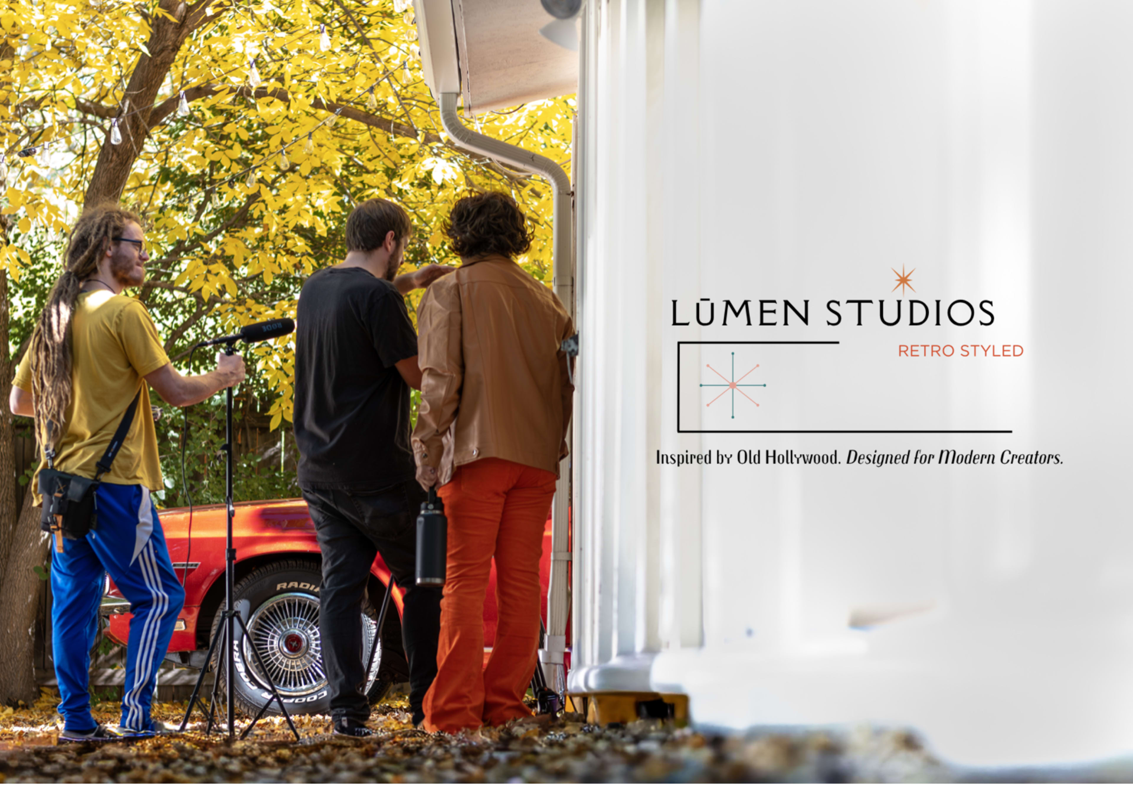 Three individuals are positioned outside a building with autumn leaves, preparing equipment near a red car.