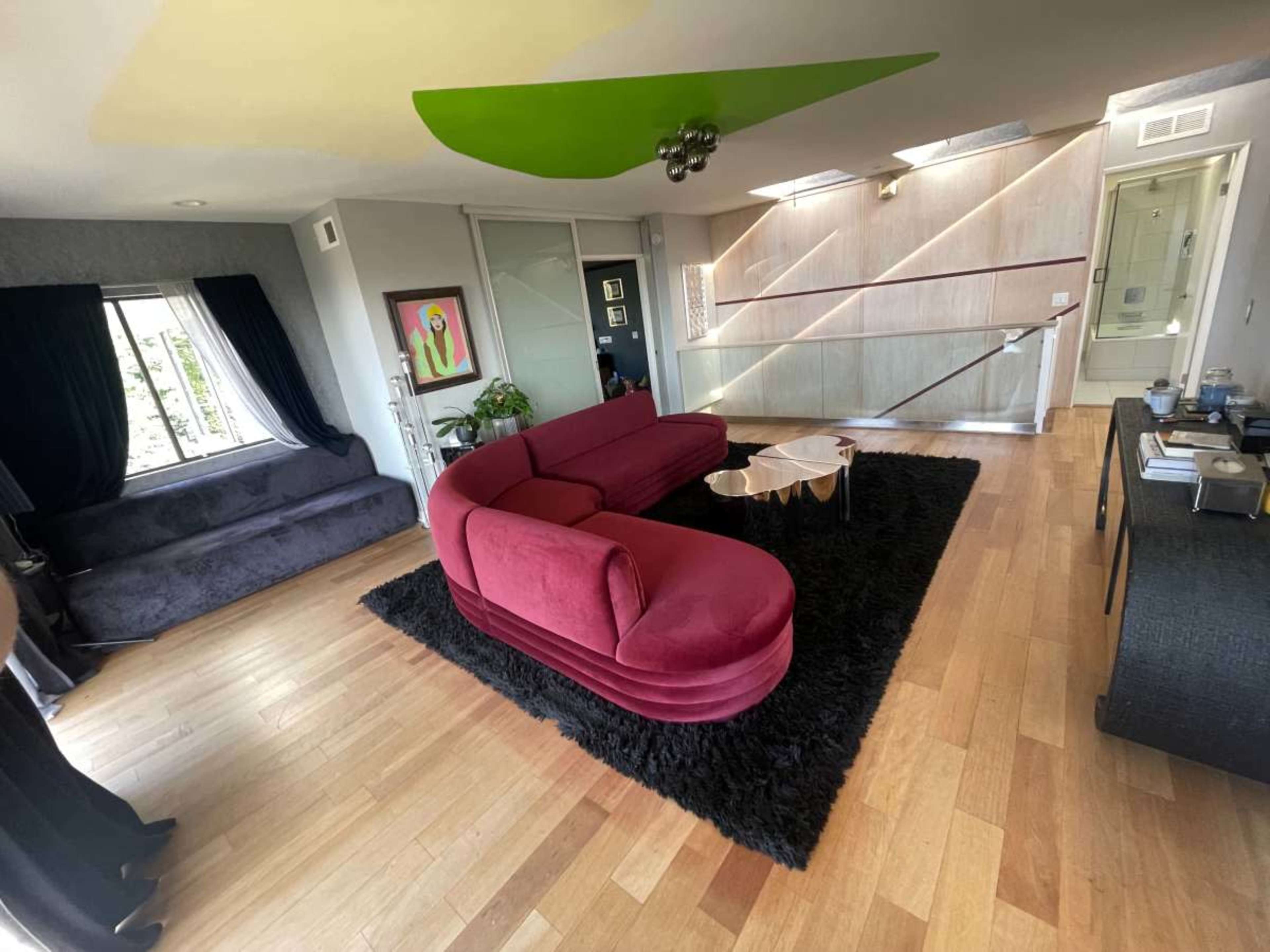 The image shows a modern living room with a large pink curved sofa, a black shag rug, and a glass coffee table in an open layout featuring wooden flooring.