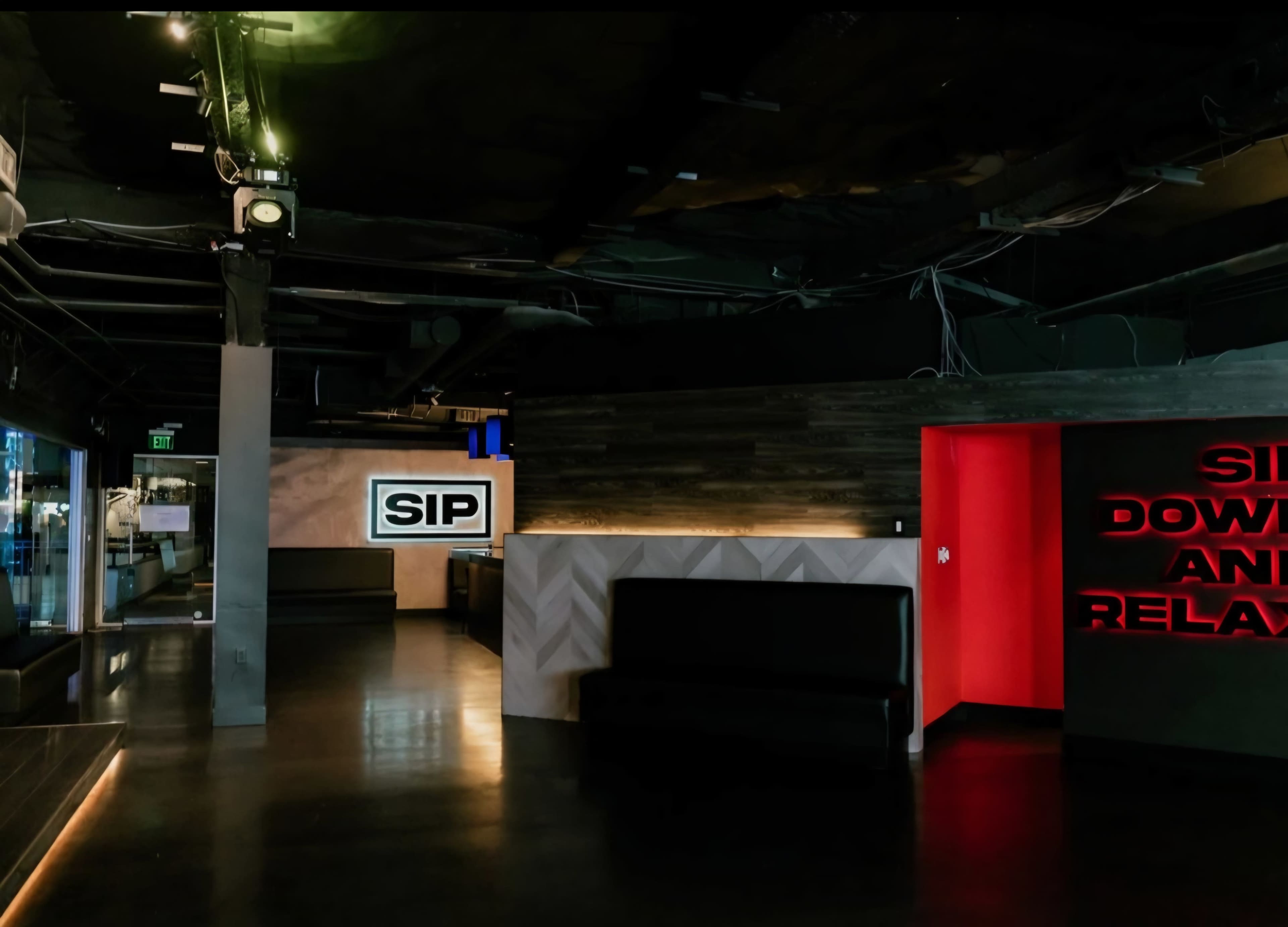 The image shows an interior of a dimly lit lounge or bar featuring a seating area, modern decor, and illuminated signage with the words "SIP" and "SIP DOWN AND RELAX."