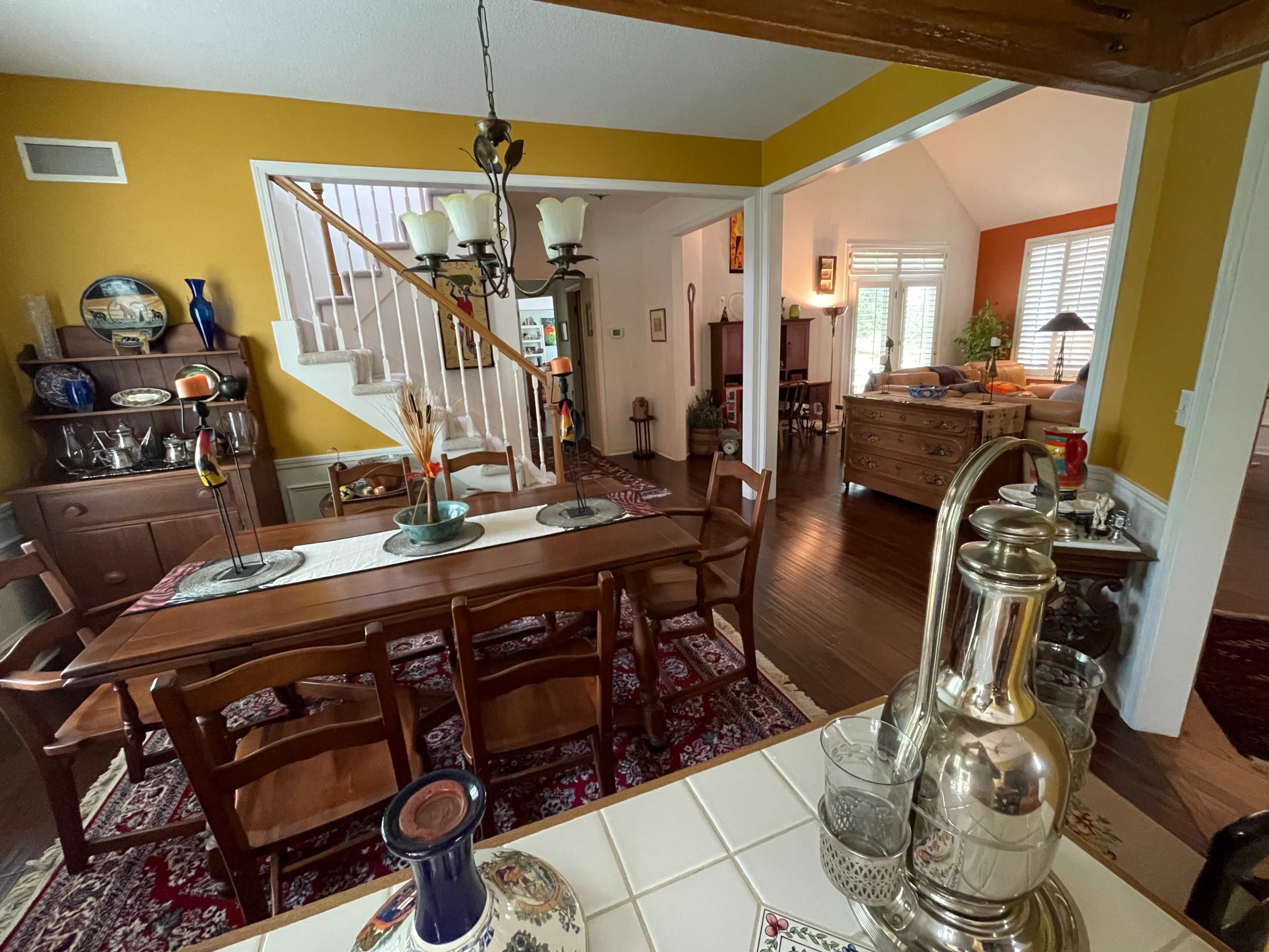The image shows a dining room with a wooden table set for a meal, adjacent to a staircase and an open living space in the background.