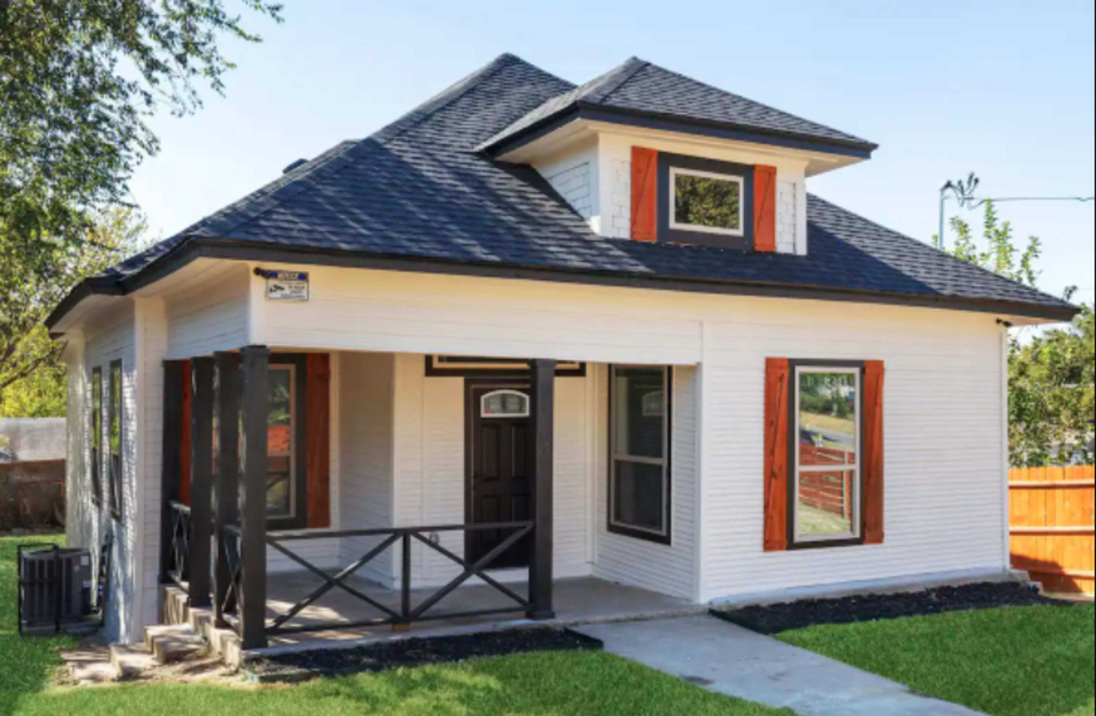 A single-story, modern-style house with a white exterior, black roof, and orange accents on the windows.
