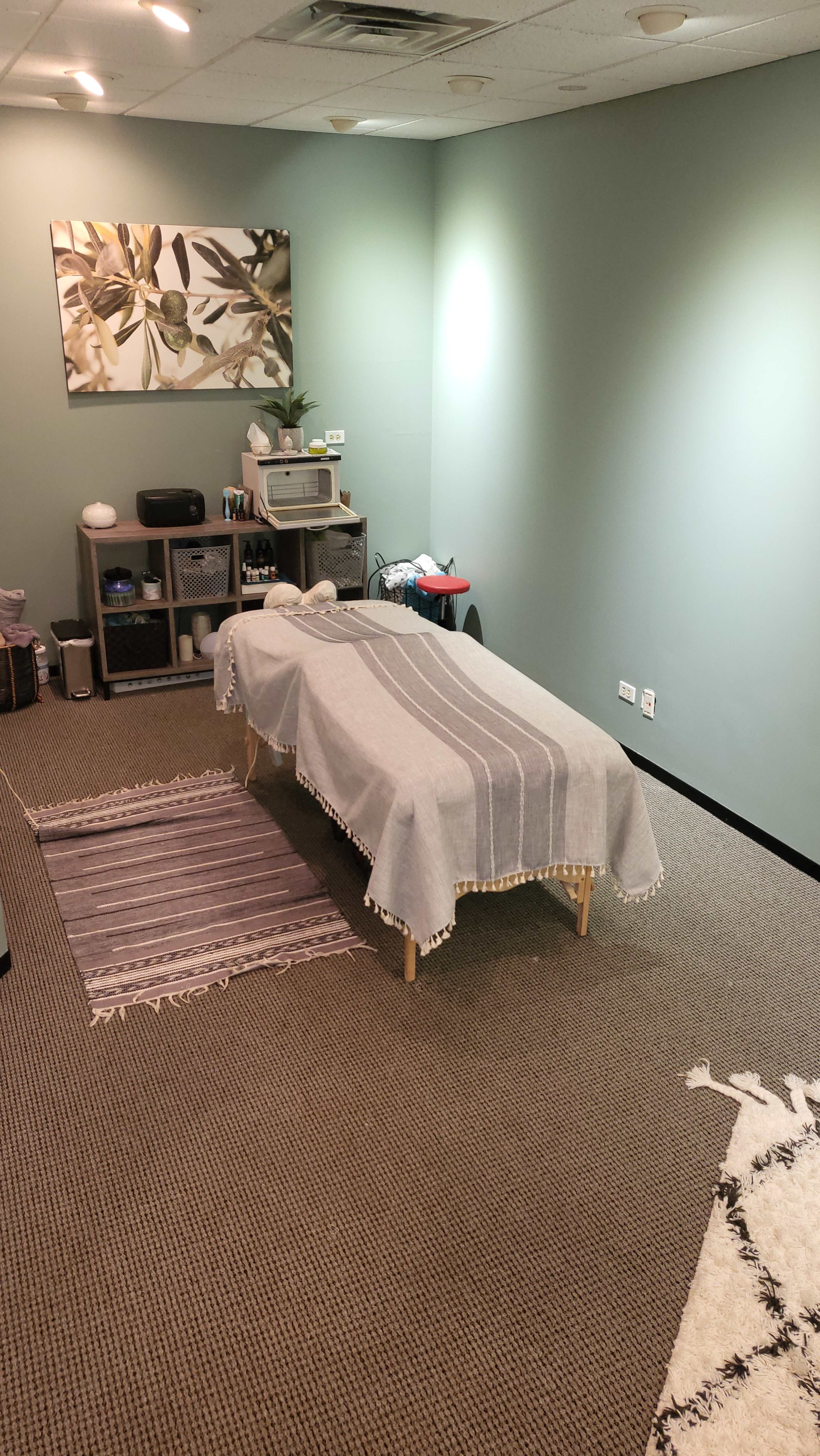 The image shows a massage therapy room featuring a massage table covered with a gray blanket, surrounded by rugs and green walls.