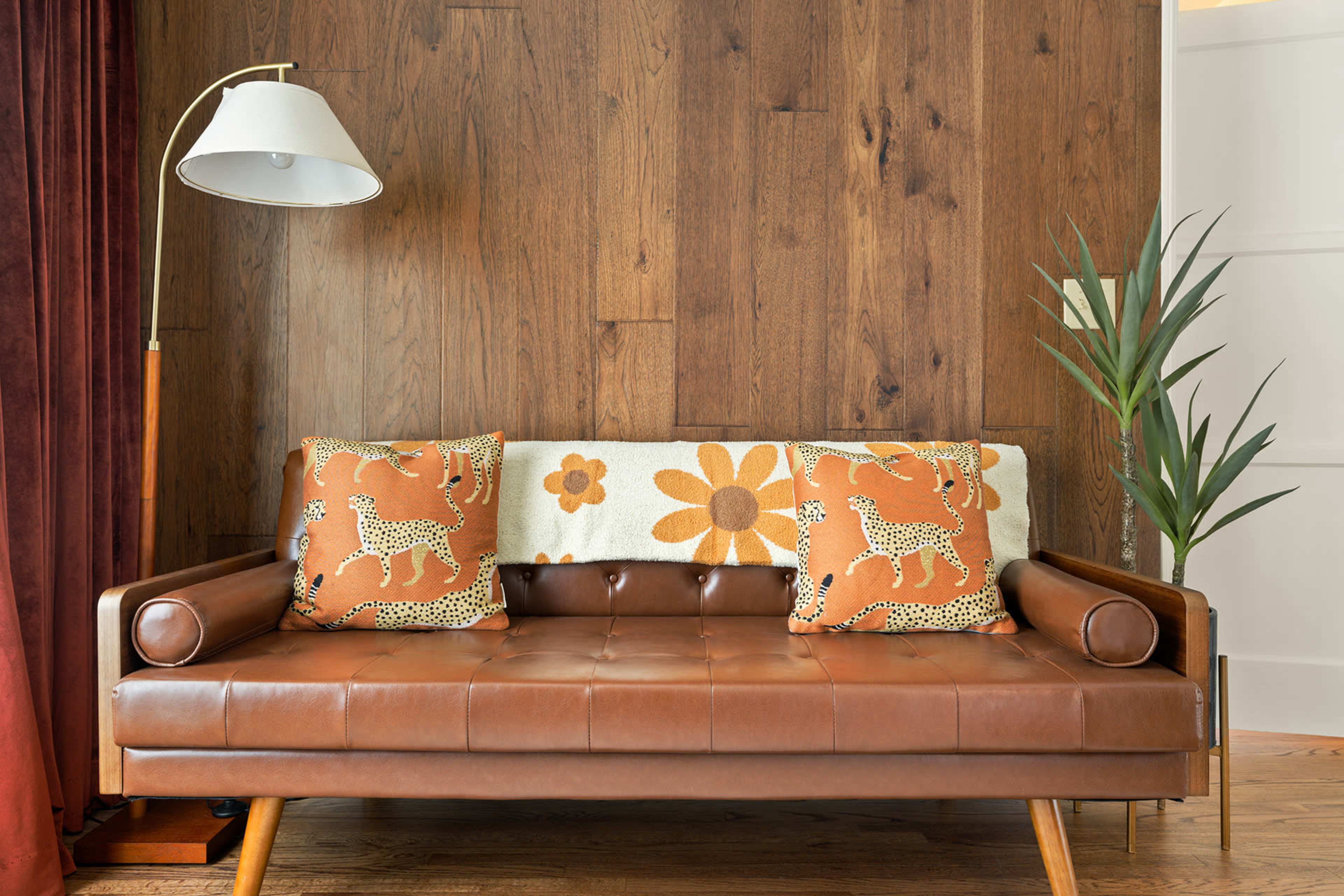 The image shows a brown leather sofa with patterned throw pillows against a wooden wall, accompanied by a floor lamp and a potted plant.