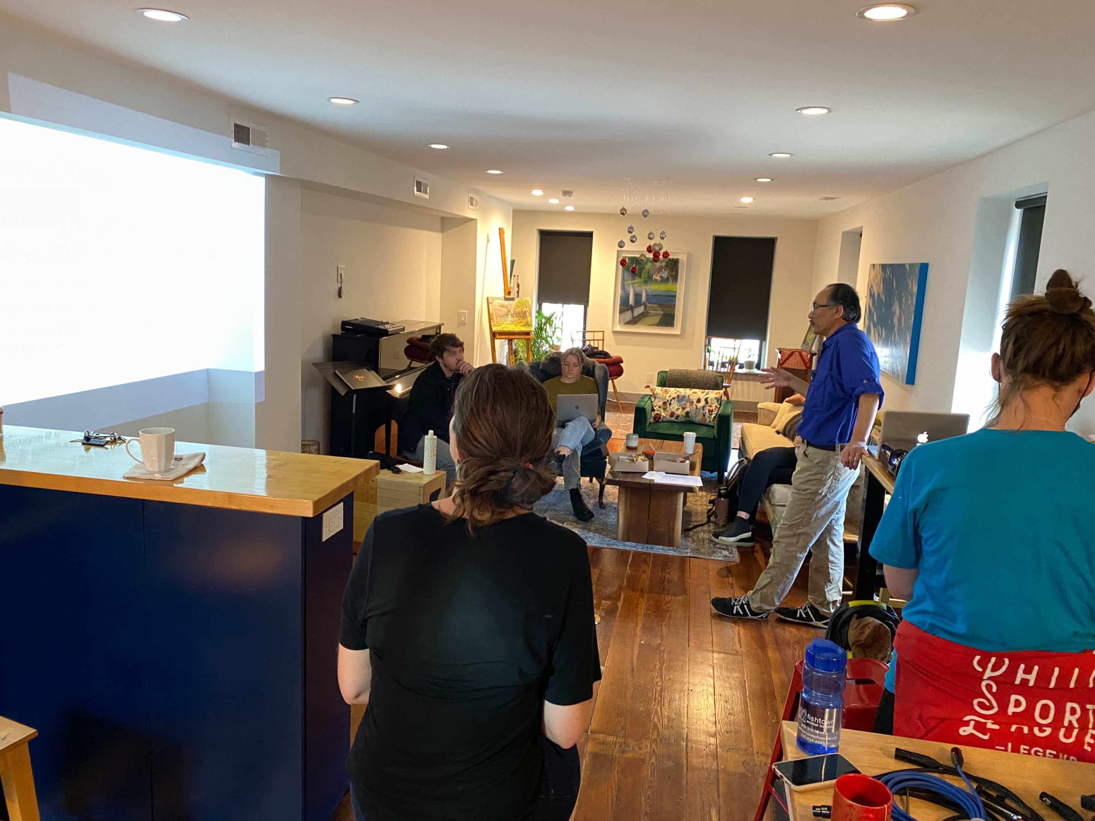 A group of people is sitting in a living room while one person stands and speaks to them, with a presentation projected on a wall.