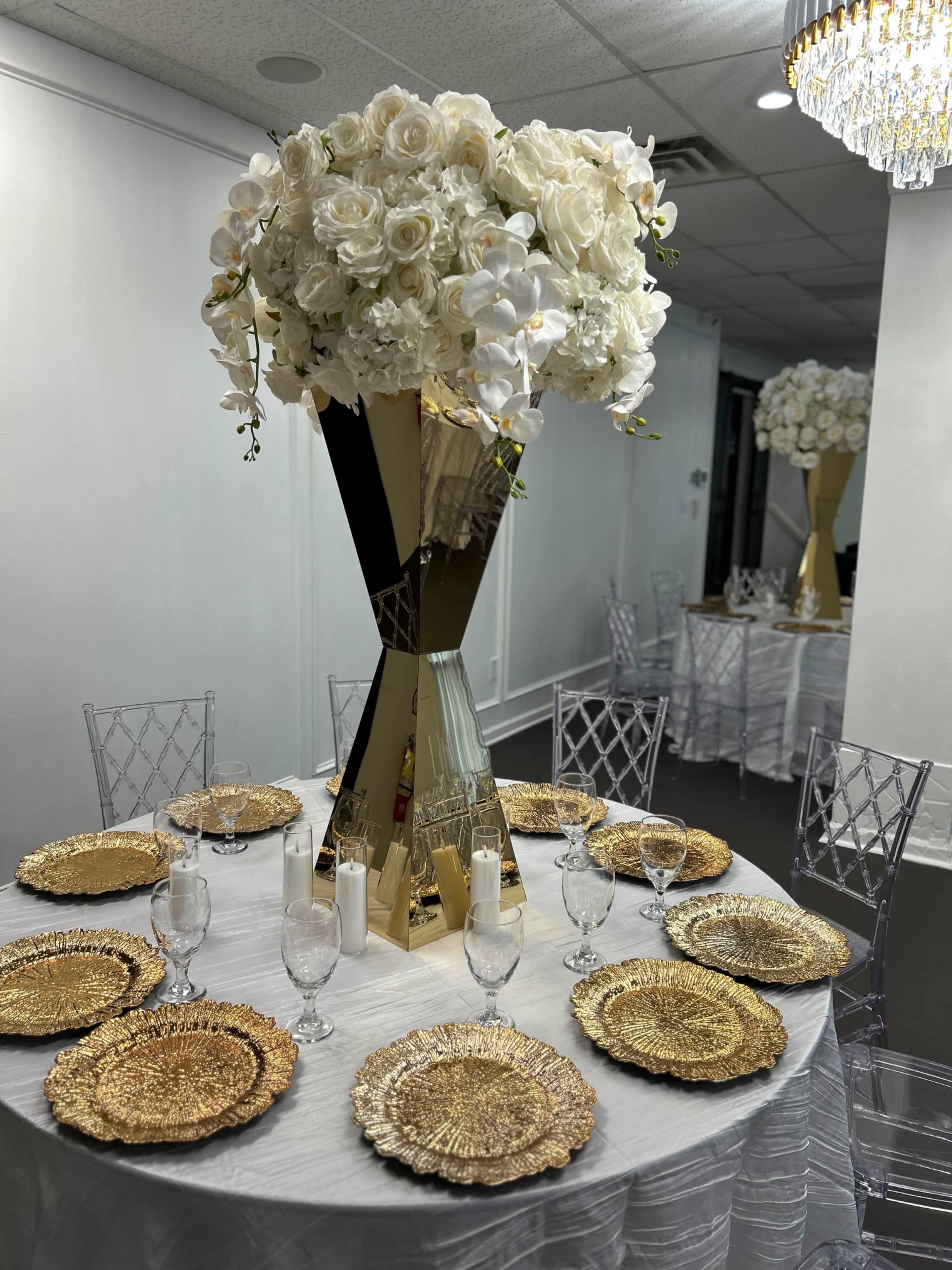 A lavish table setting features a tall floral arrangement of white roses and orchids on a gold pedestal, surrounded by gold-rimmed plates and clear glassware.