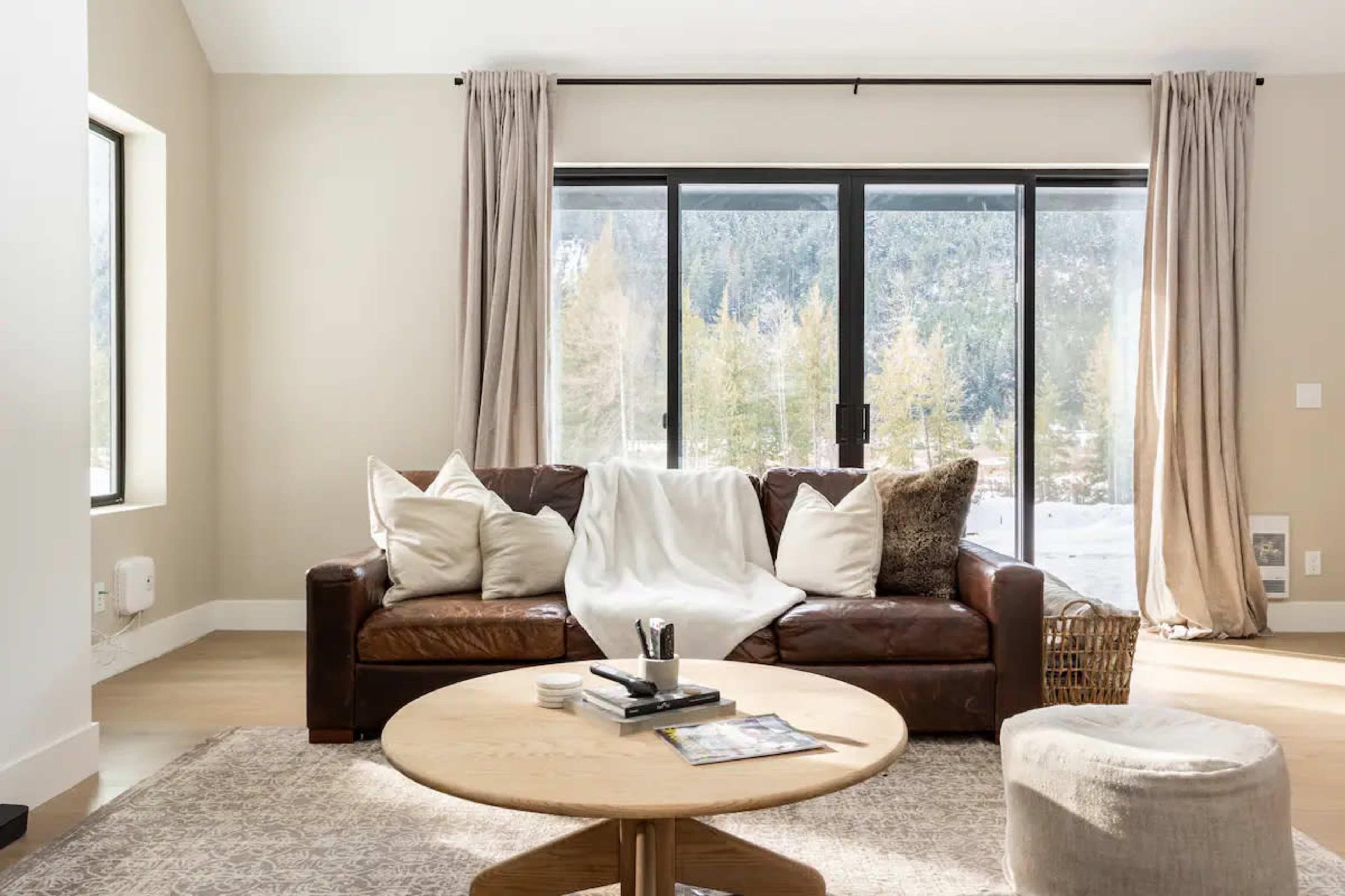A leather sofa with pillows and a blanket sits in front of large windows overlooking a snowy landscape, accompanied by a round wooden coffee table and a small ottoman.