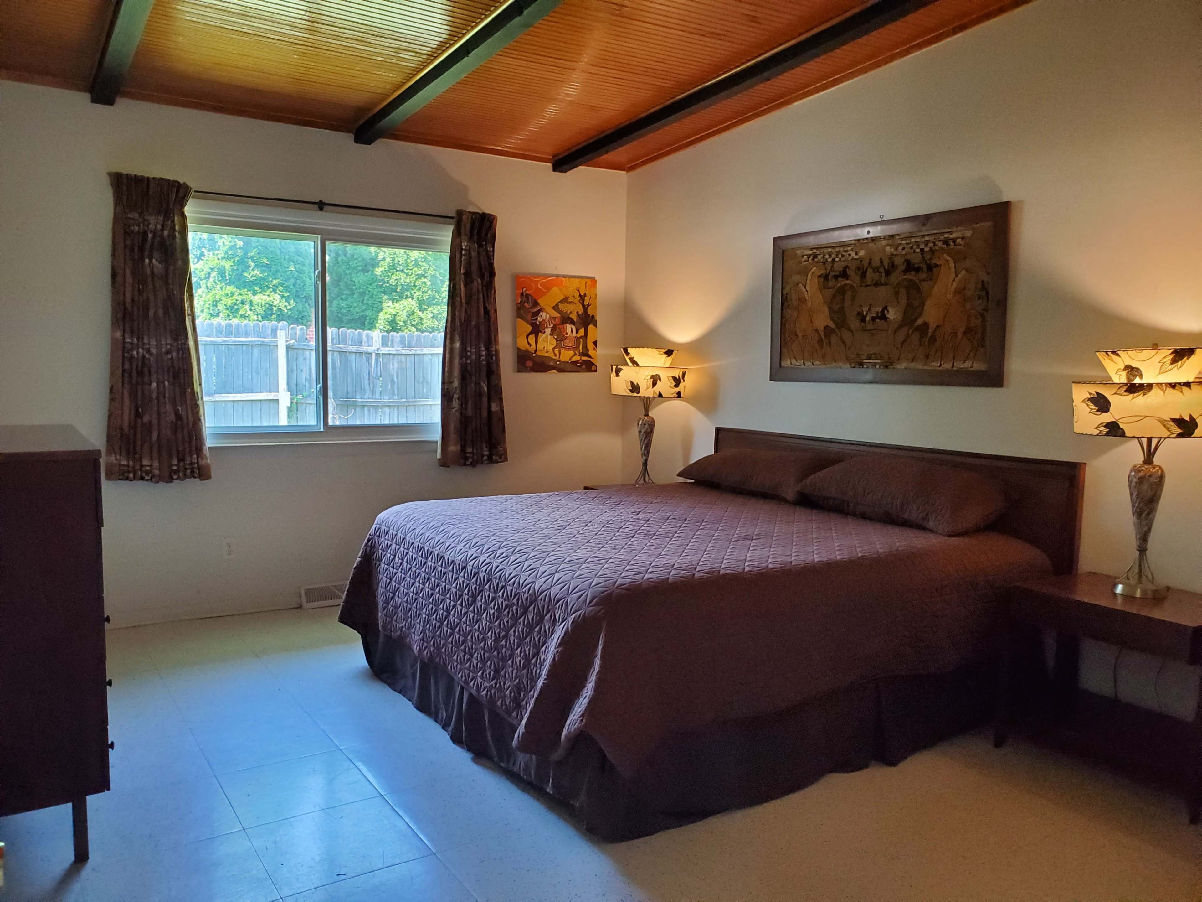 A simple bedroom with a queen-sized bed, two bedside lamps, a window, and artwork on the walls.