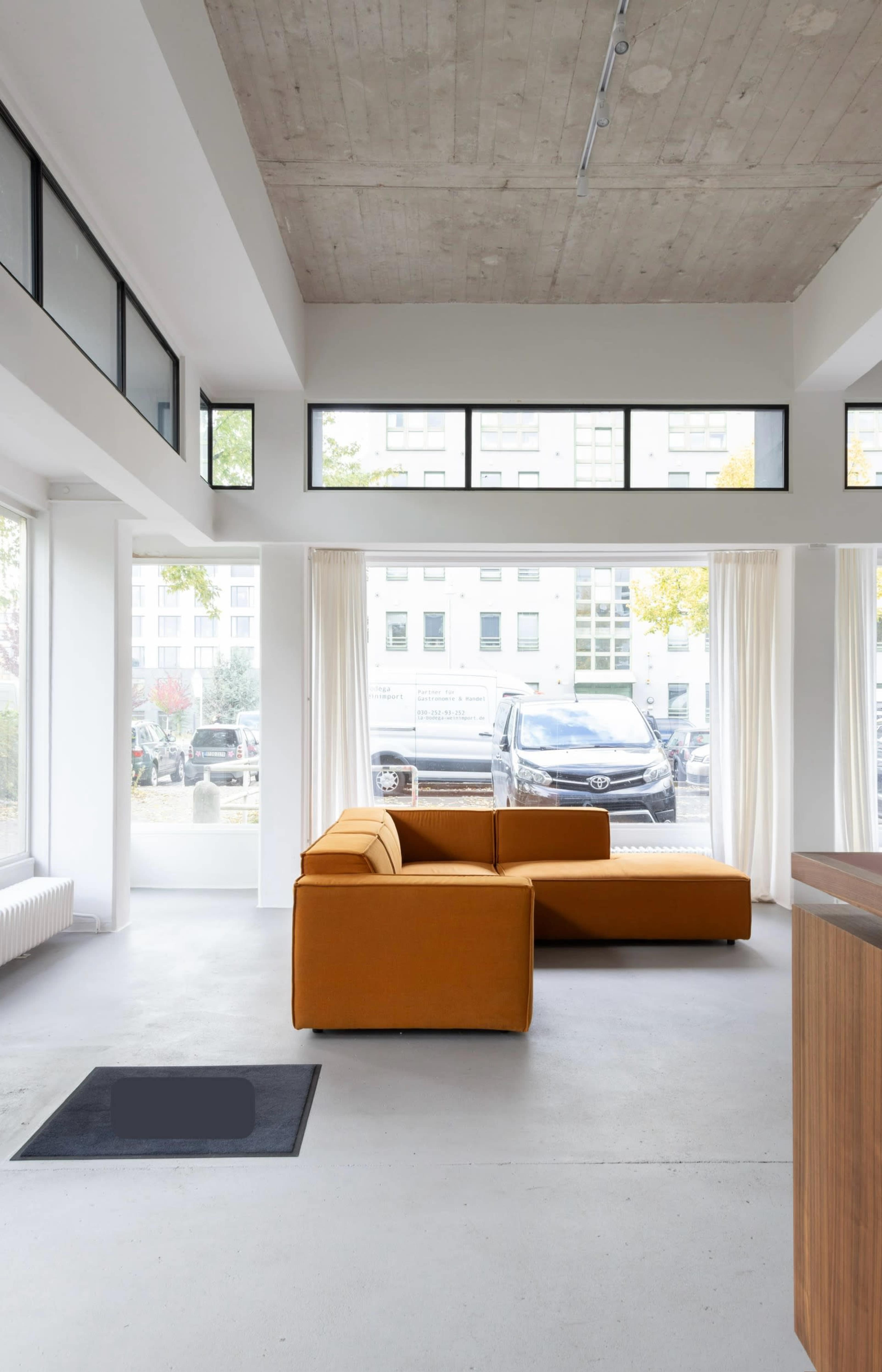 A modern living room features a brown sectional sofa, large windows providing natural light, and minimalist decor.