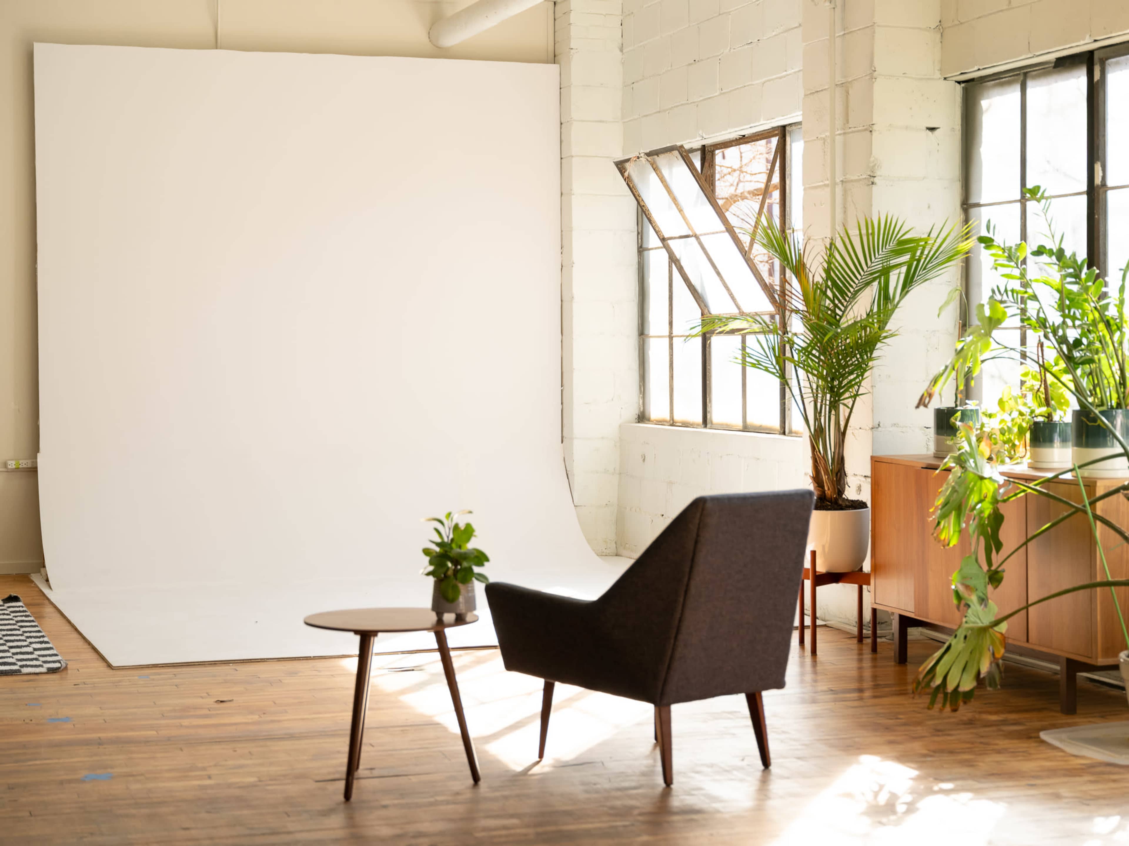 The image features a minimalist interior with a large white backdrop, a dark chair, a round table with a plant, and a tall window allowing natural light to fill the space.