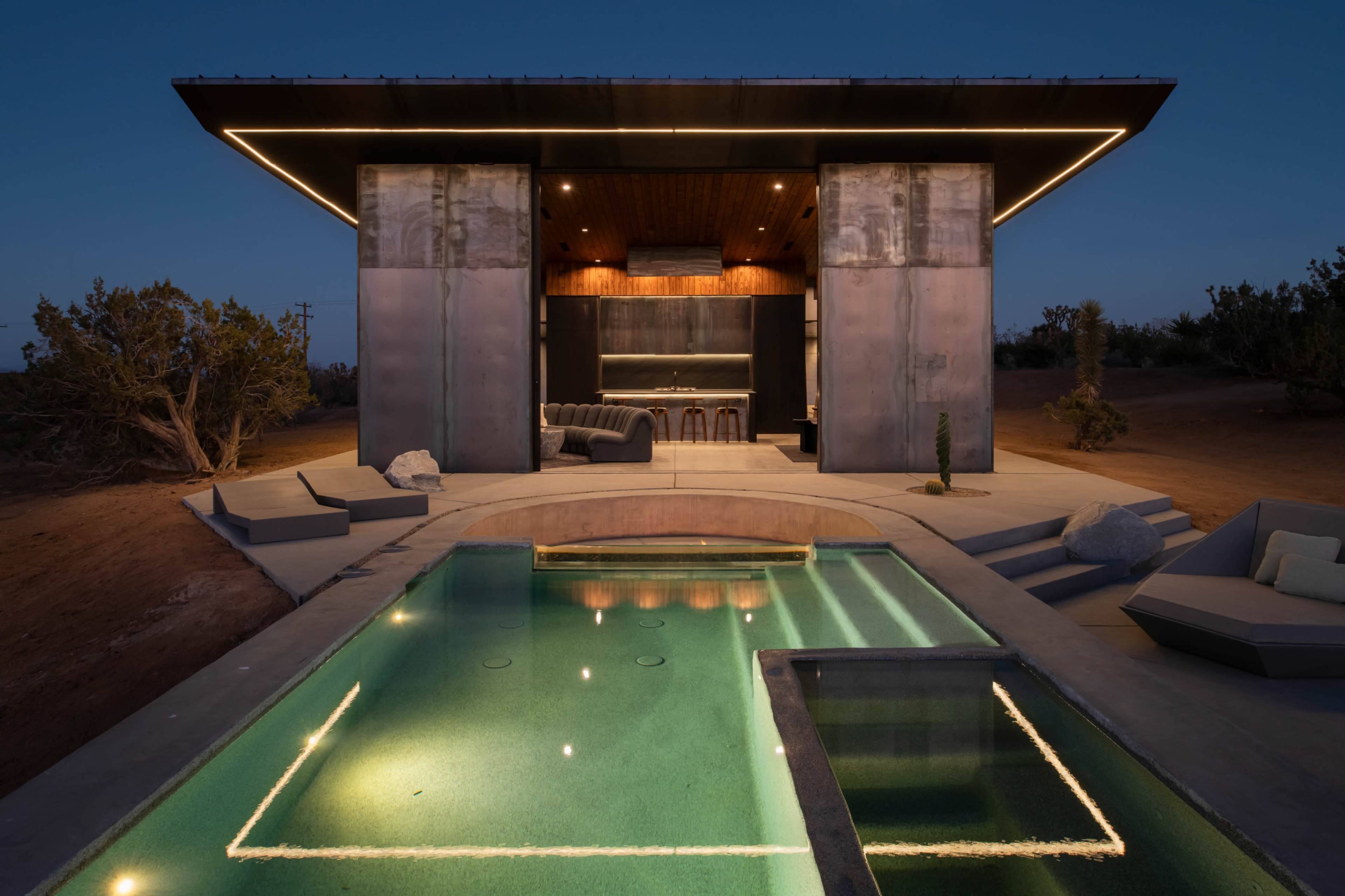 A modern outdoor space features a sleek pool area in front of a minimalist structure with a kitchen and seating area, illuminated by warm lighting at dusk.