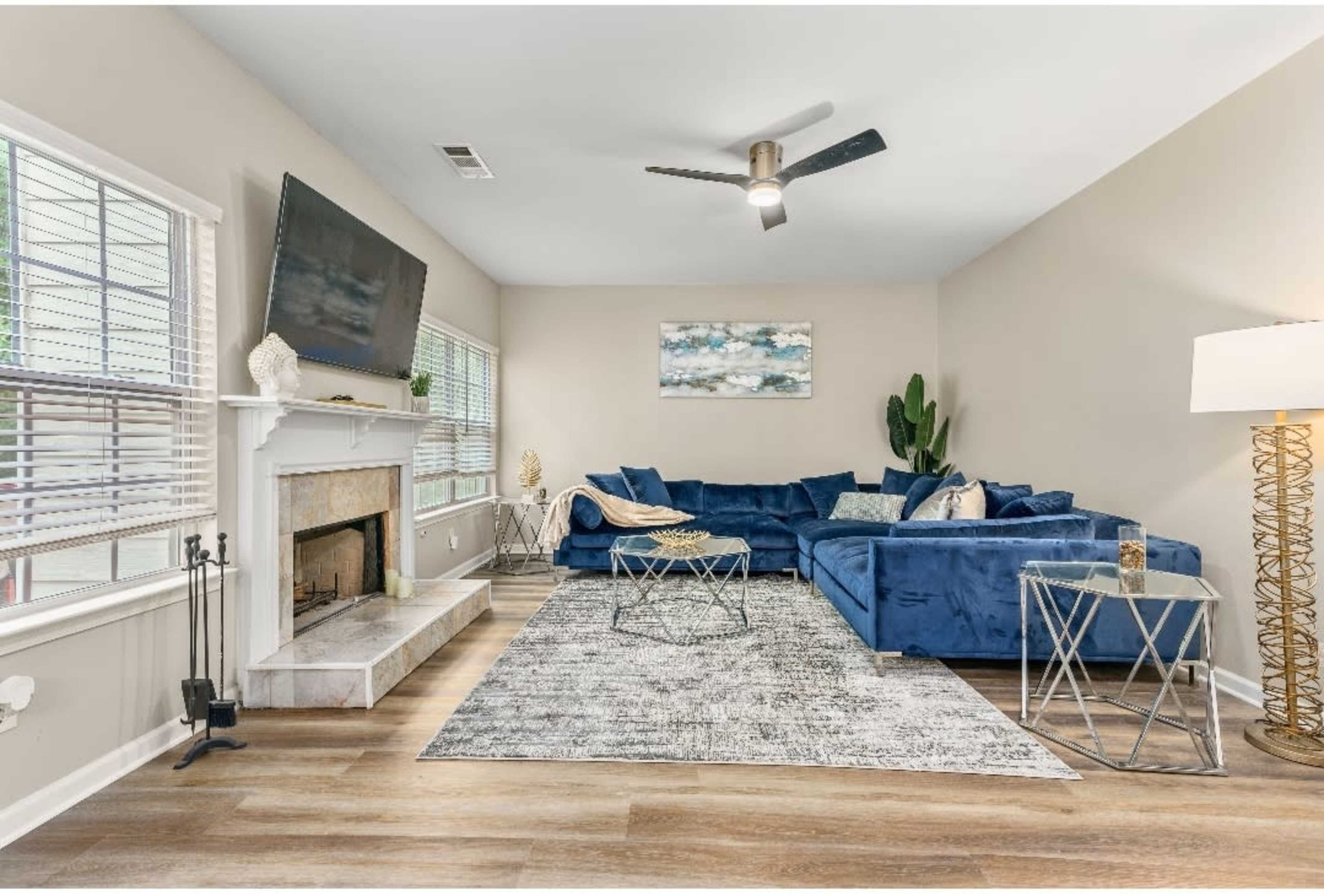 A spacious living room featuring a blue sectional couch, a modern ceiling fan, a fireplace with a TV above it, and a stylish coffee table on a patterned area rug.