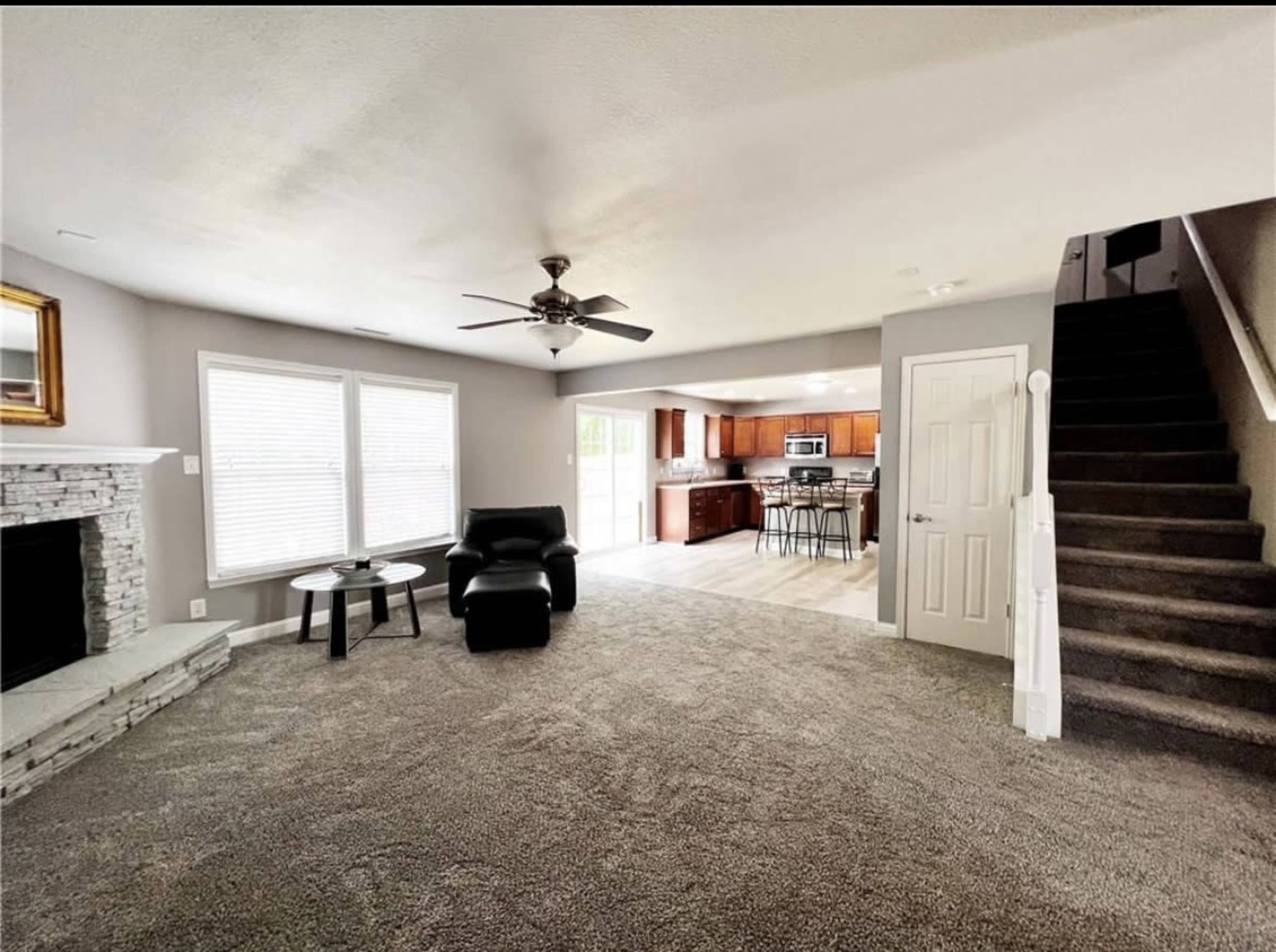A spacious living room with a fireplace and carpet, adjacent to a staircase and a kitchen area in the background.