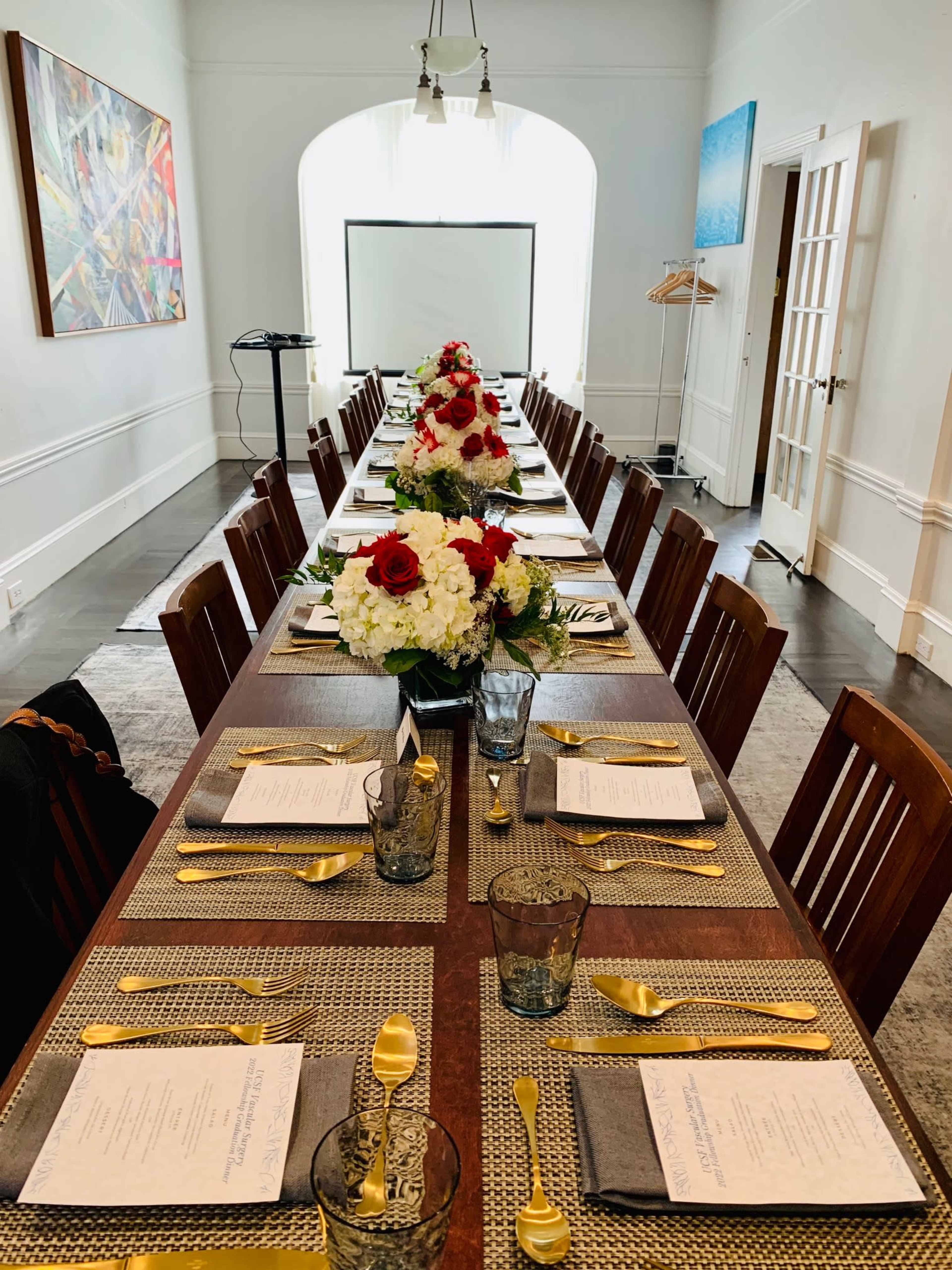 A long dining table is set with elegant place settings and floral centerpieces, arranged in a brightly lit room with a projector screen in the background.