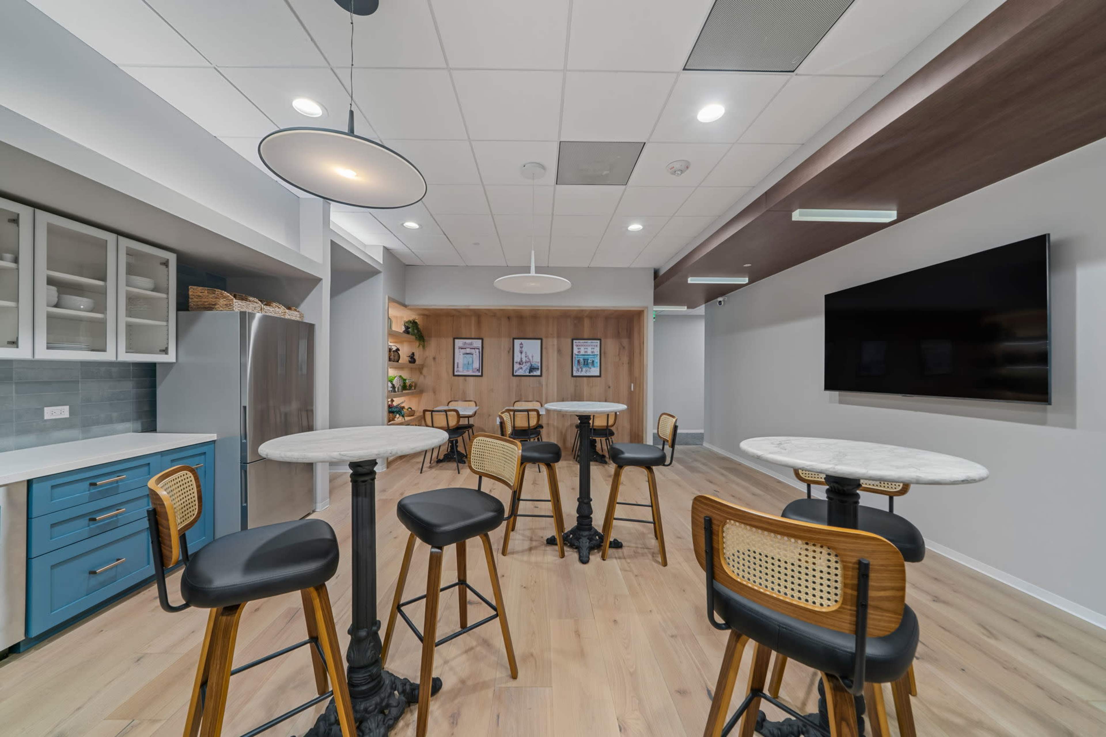 The image shows a modern lounge area with high tables, stools, a kitchenette, and a large screen on the wall.