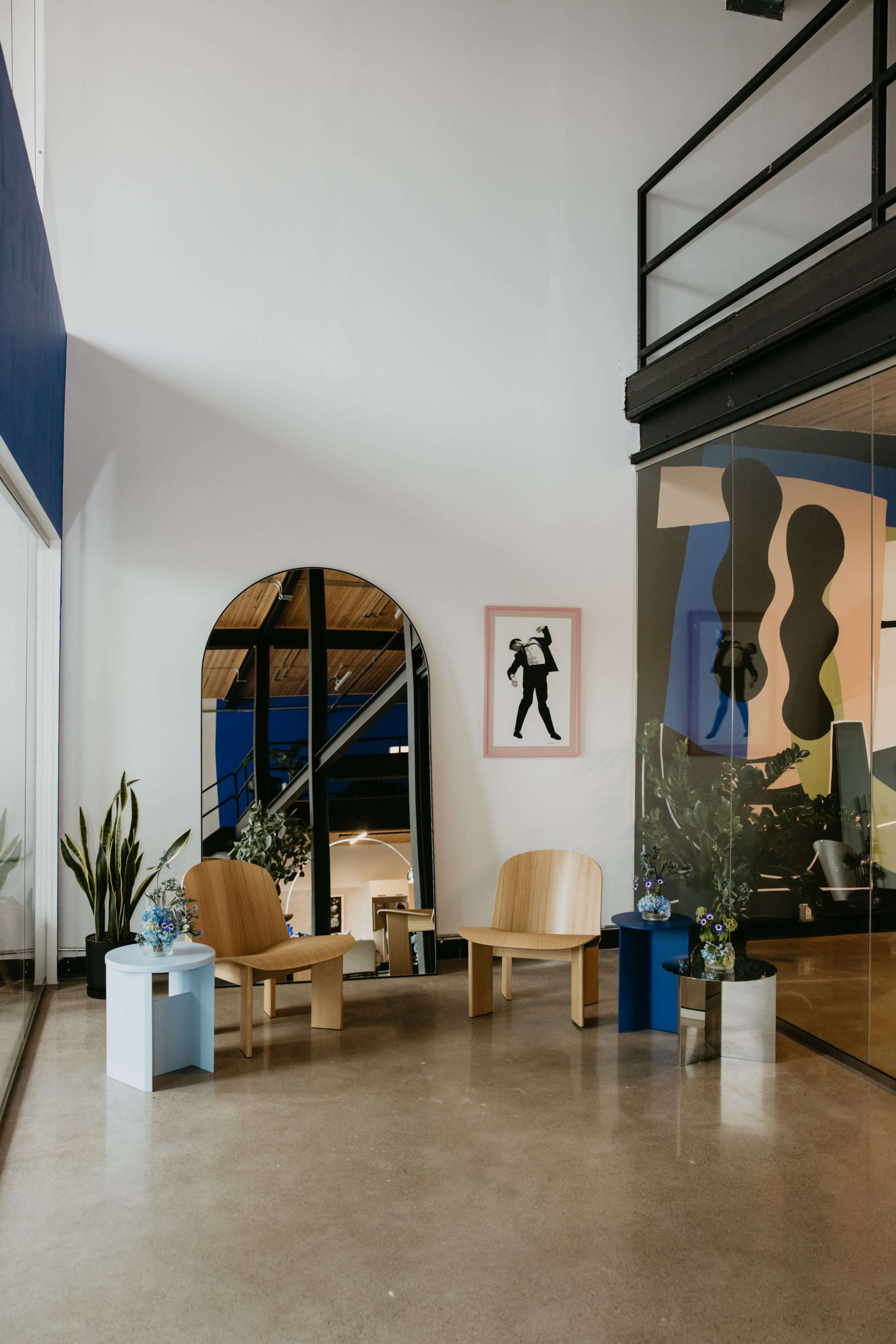 The image shows a modern interior space featuring two wooden chairs, a small table, and decorative plants, with large windows and a vibrant blue accent wall.