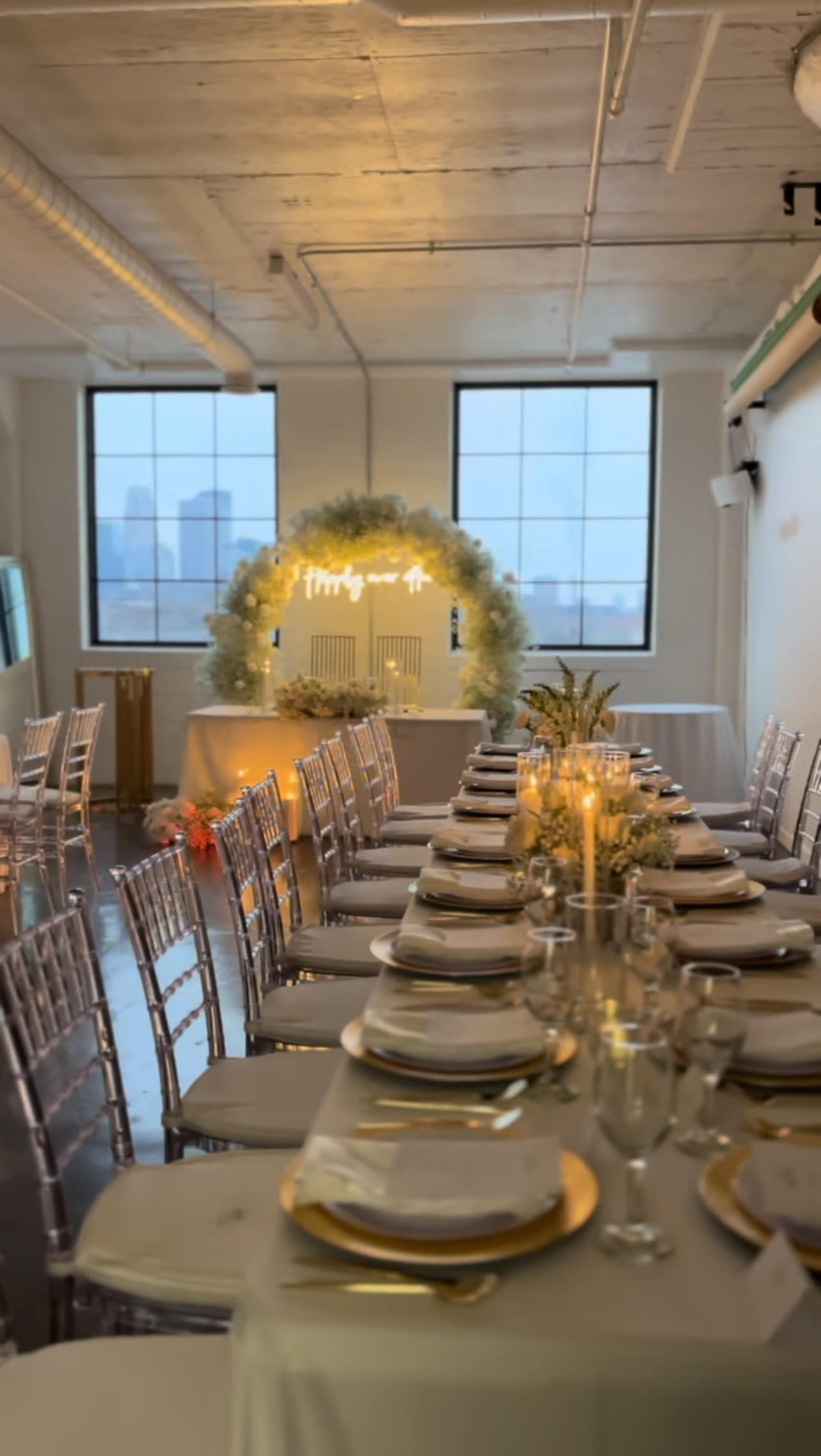 A long banquet table is set with elegant tableware in a well-lit room featuring large windows overlooking a city skyline.
