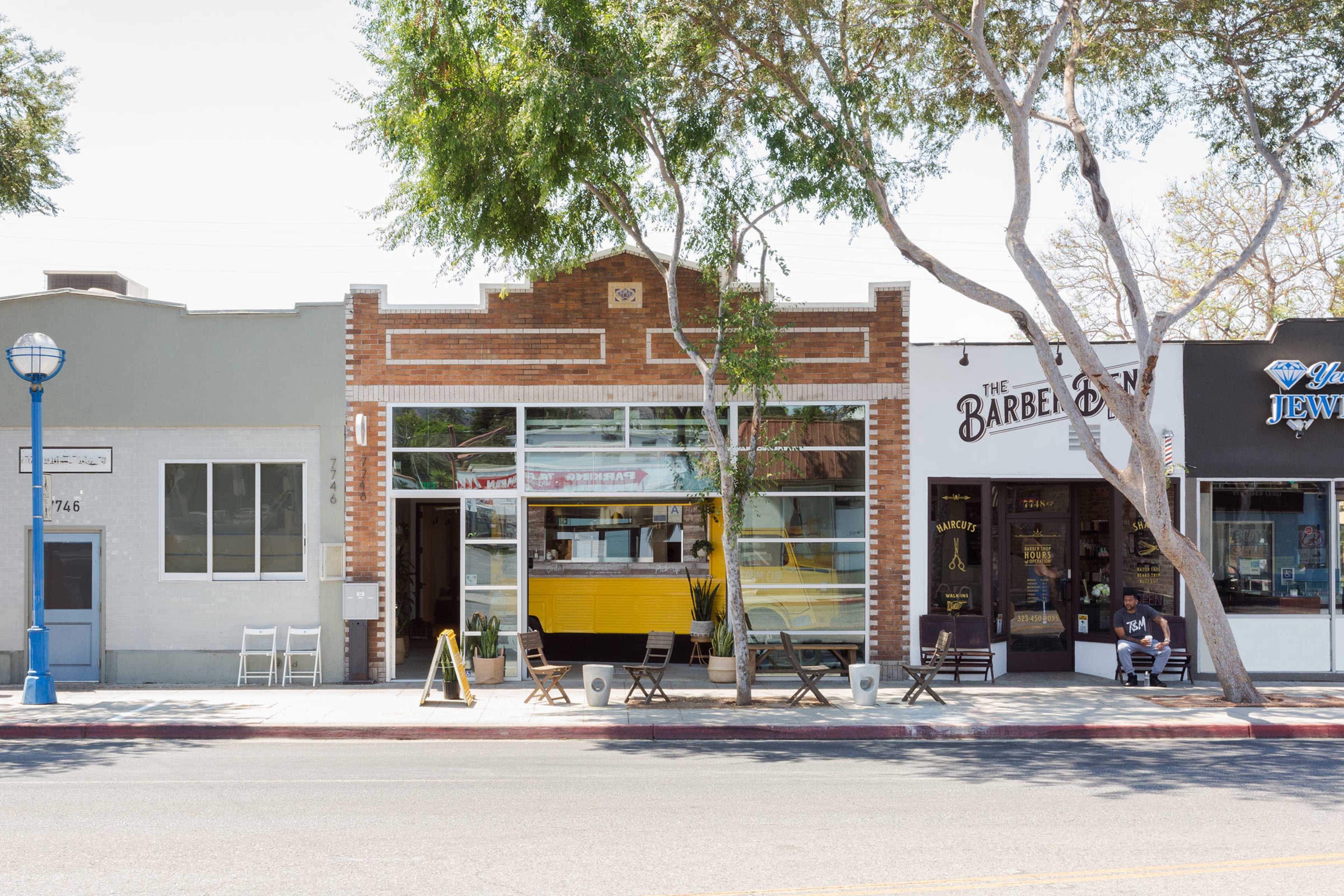 The image shows a street view featuring a brick building with large windows, a yellow door, and seating arrangements outside, alongside a barber shop and other establishments.