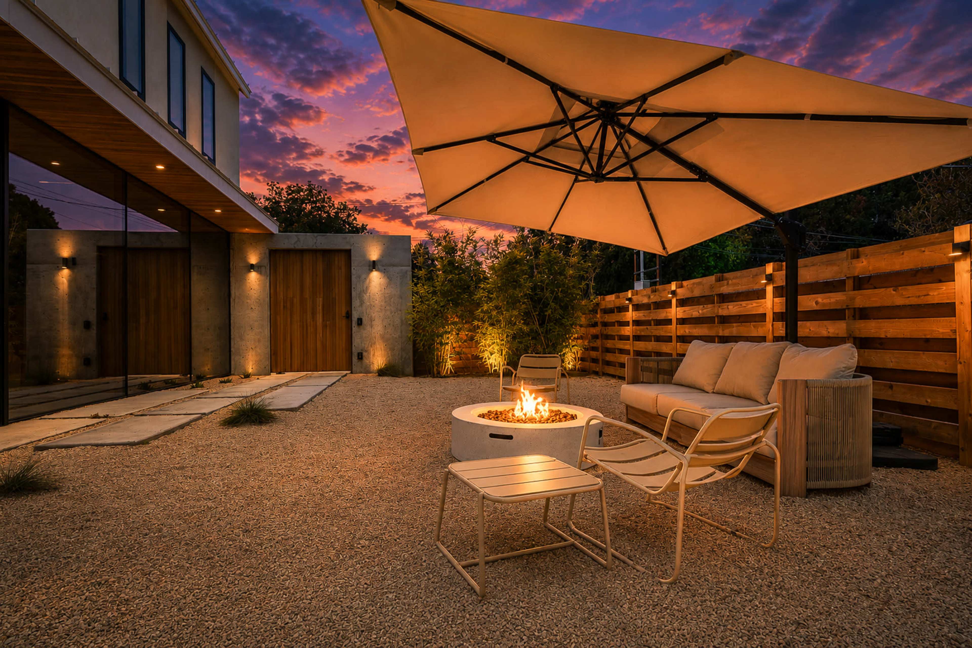 A modern outdoor seating area features a fire pit surrounded by a sofa and tables, with an umbrella providing shade against a colorful sunset backdrop.