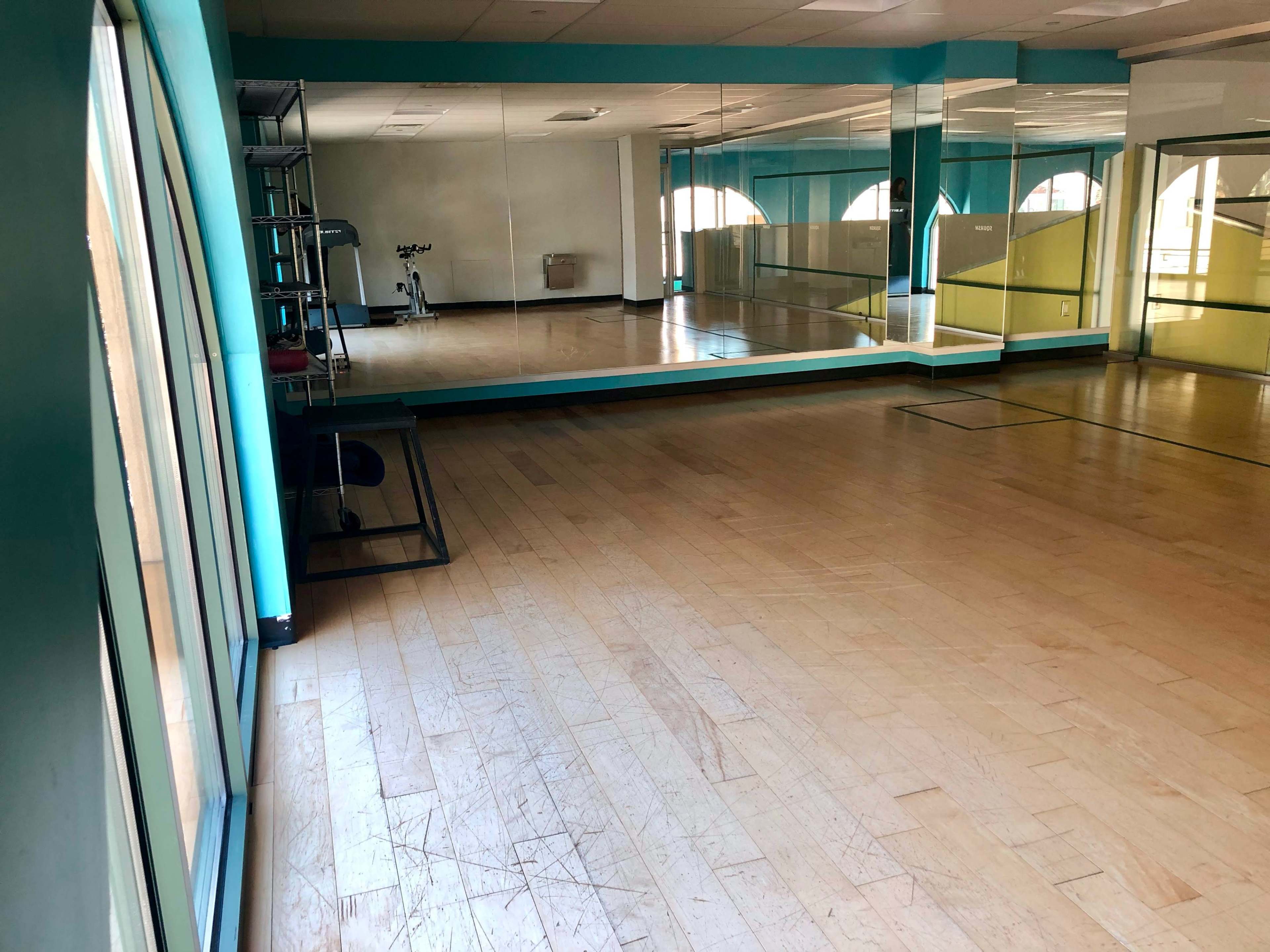 An empty fitness studio with wooden flooring and large mirrors on the walls, illuminated by natural light from windows.