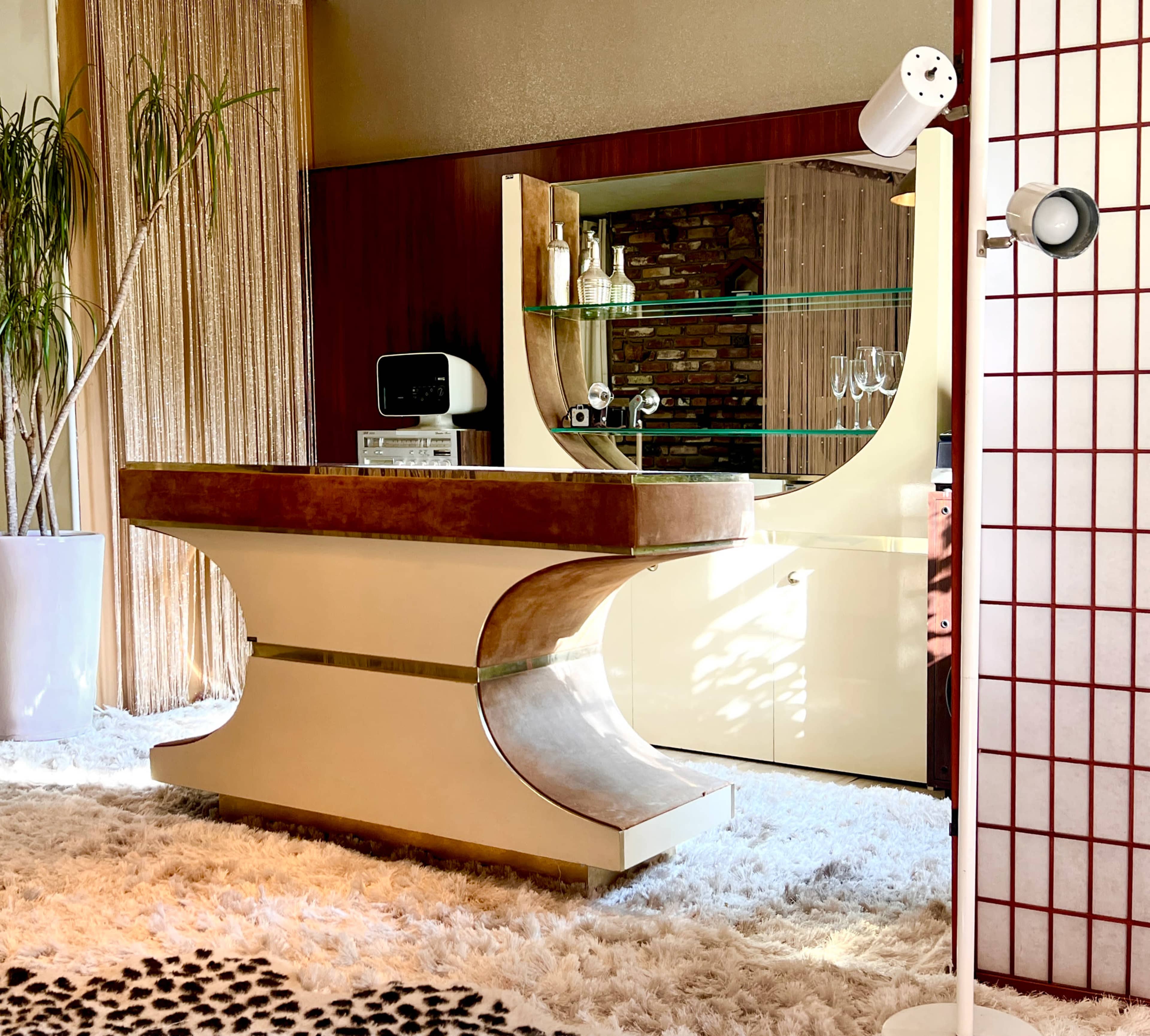 A retro-style bar counter with a curved design, surrounded by decorative elements like a potted plant, a modern lamp, and a light-colored rug.
