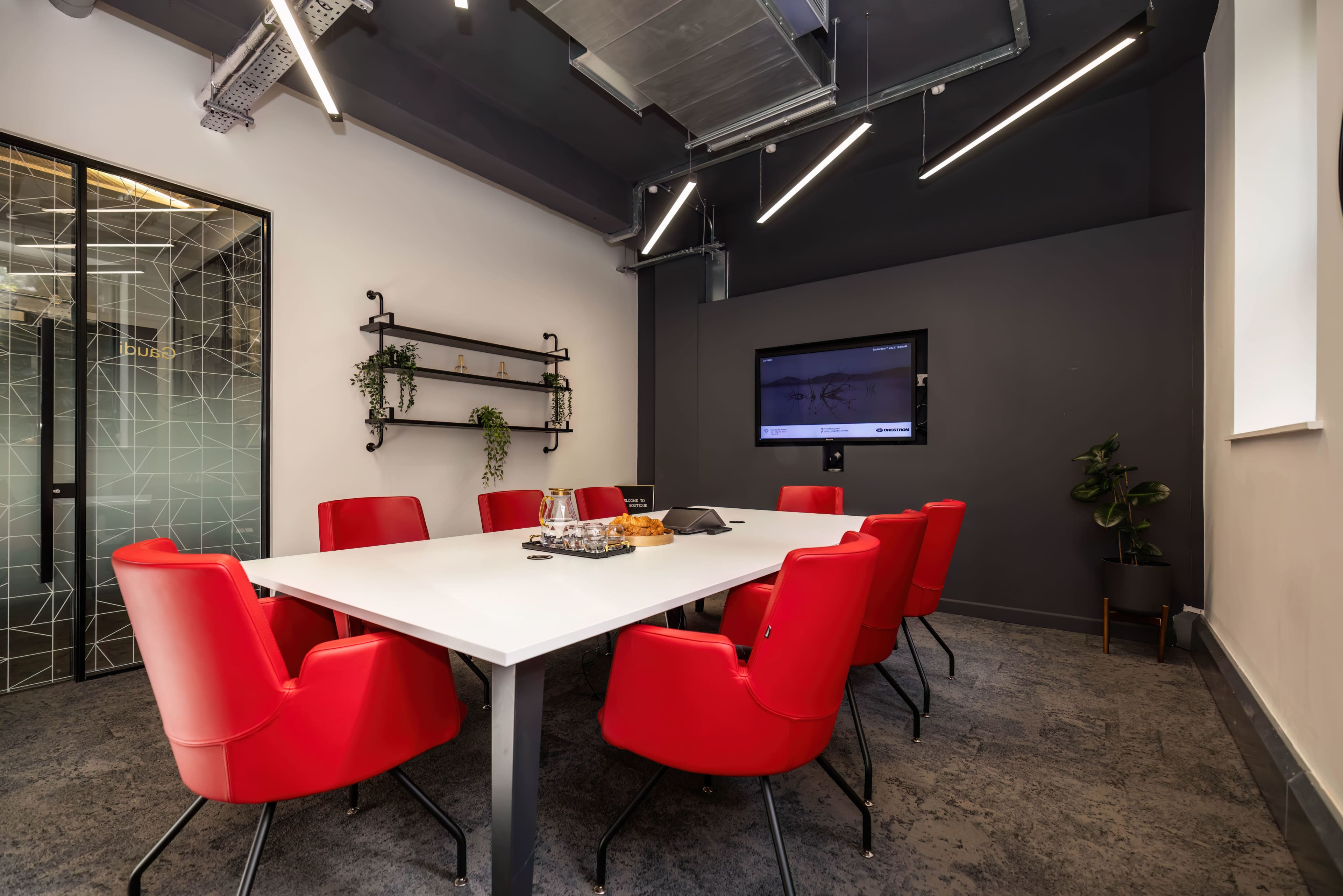 A conference room features a large white table surrounded by red chairs, with a large screen and shelves displaying plants on the walls.