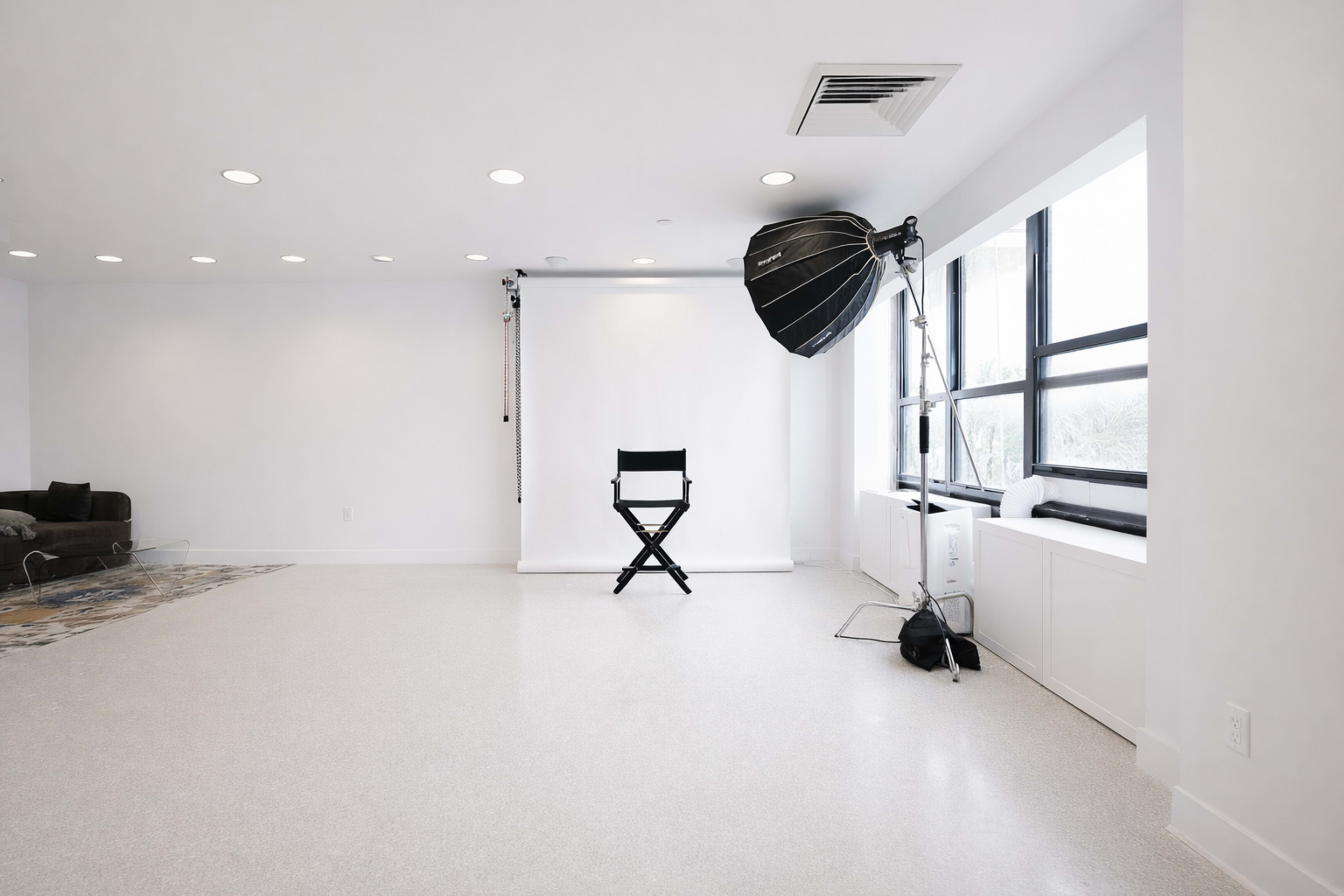 The image shows a well-lit, minimalist studio space featuring a black director's chair, a large softbox light, and a white backdrop.