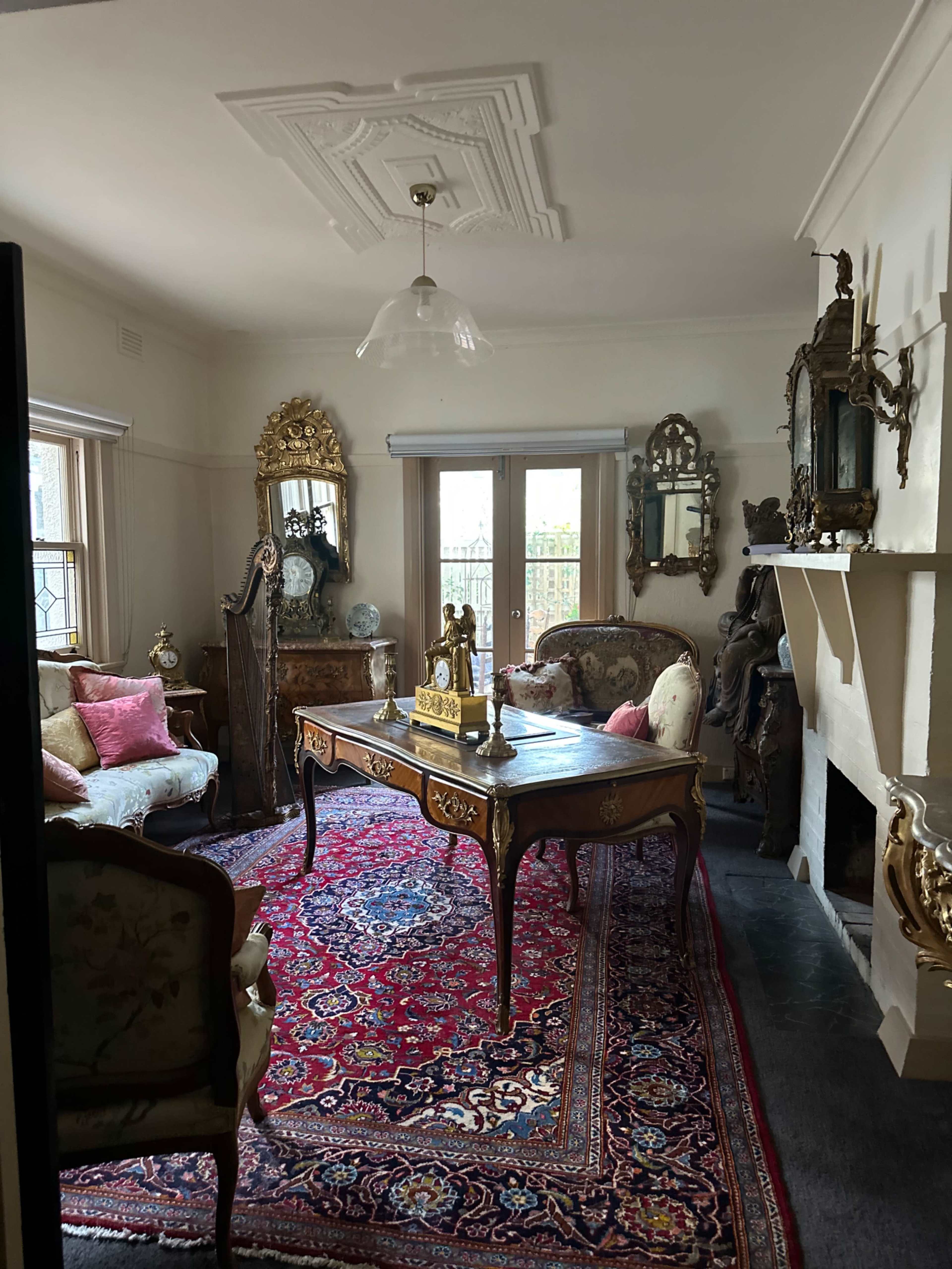 The room features ornate furniture, including a wooden table and a vintage sofa, set against an intricately patterned rug and decorated with mirrors and sculptures.