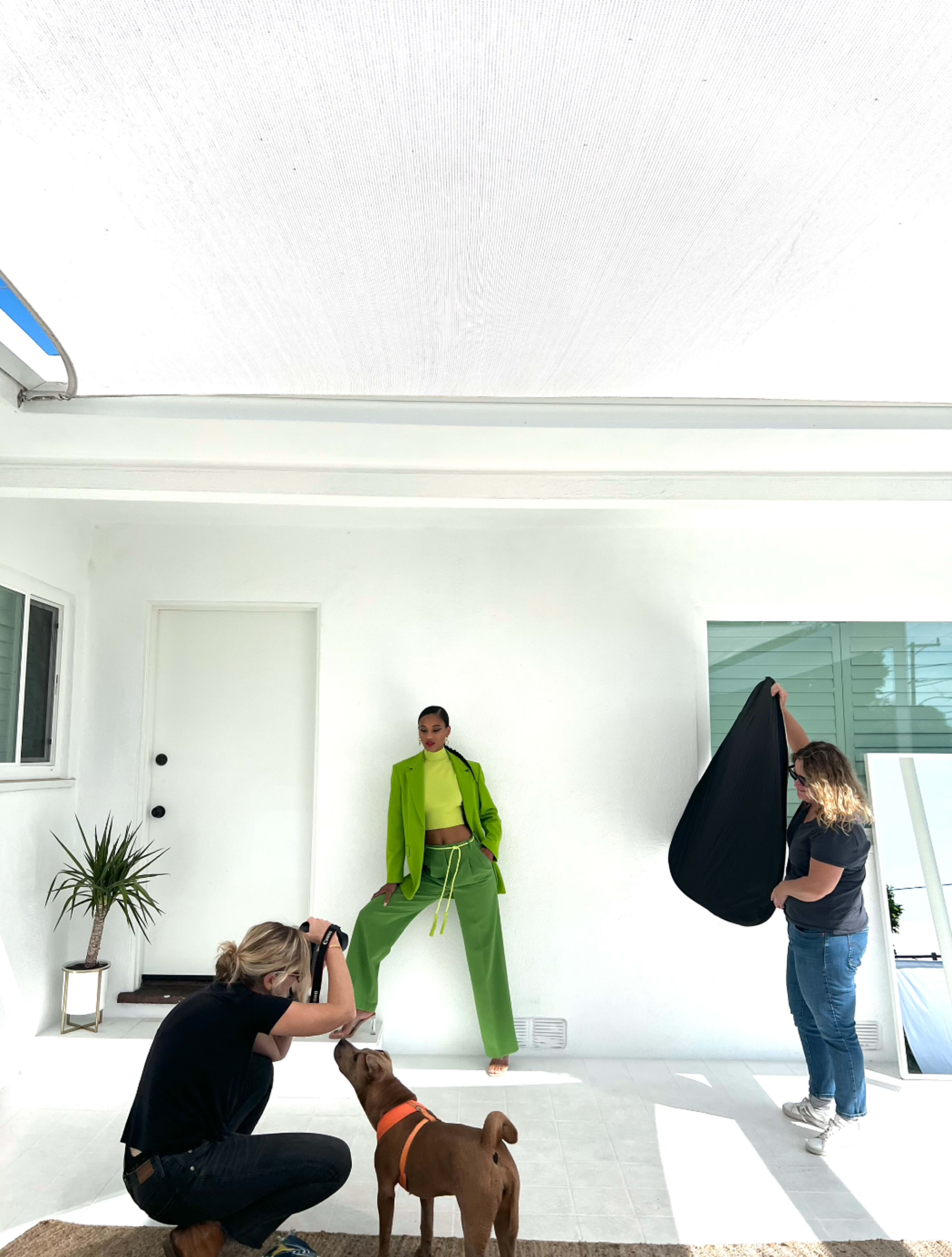 A model in a bright green outfit poses against a white wall while two people assist with the shoot and a dog looks on.