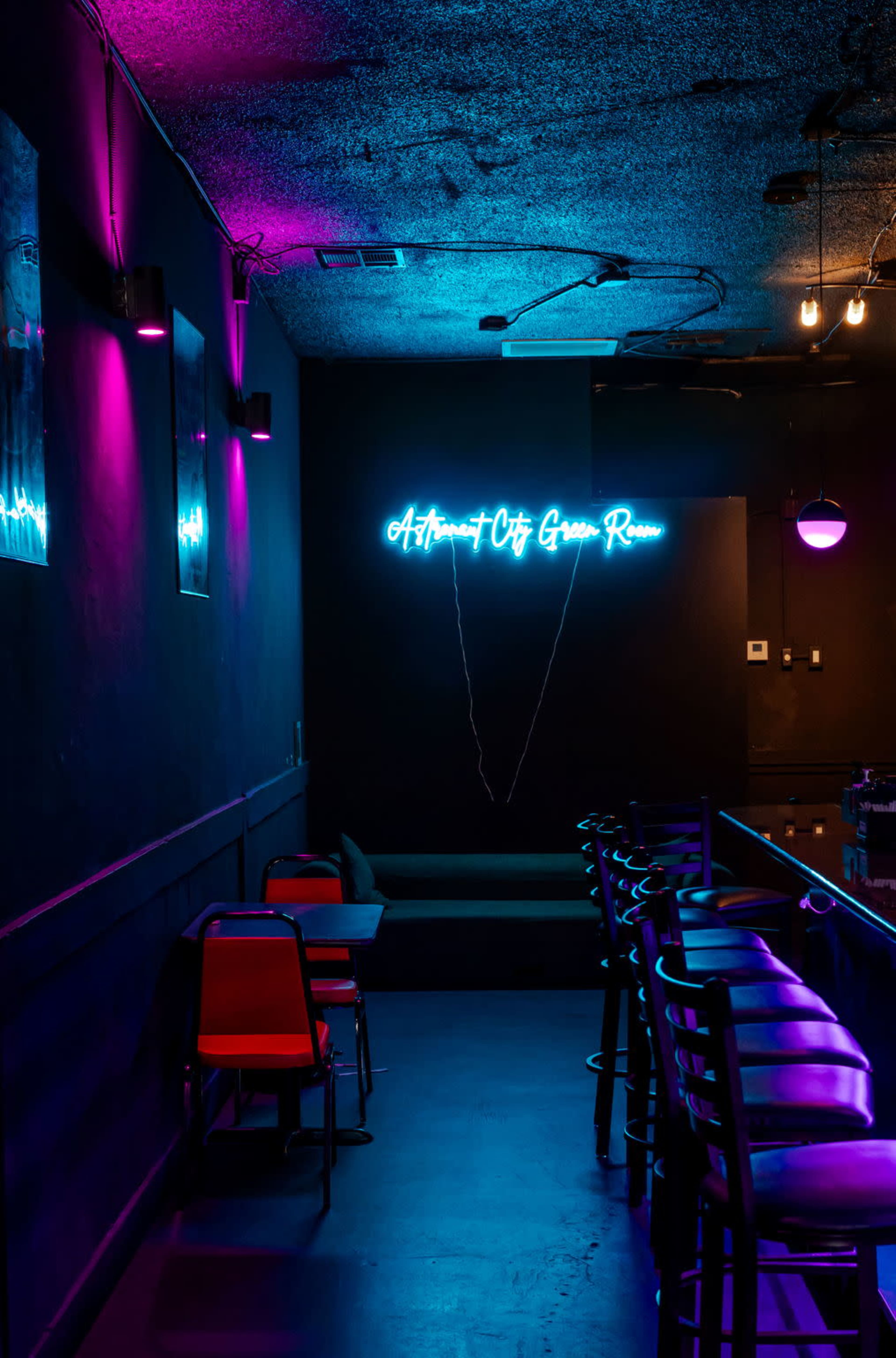 The image shows a dimly lit bar area featuring neon signage that reads "Artisan City Game Room," with a row of empty chairs and a dark wall adorned with purple and blue lighting.