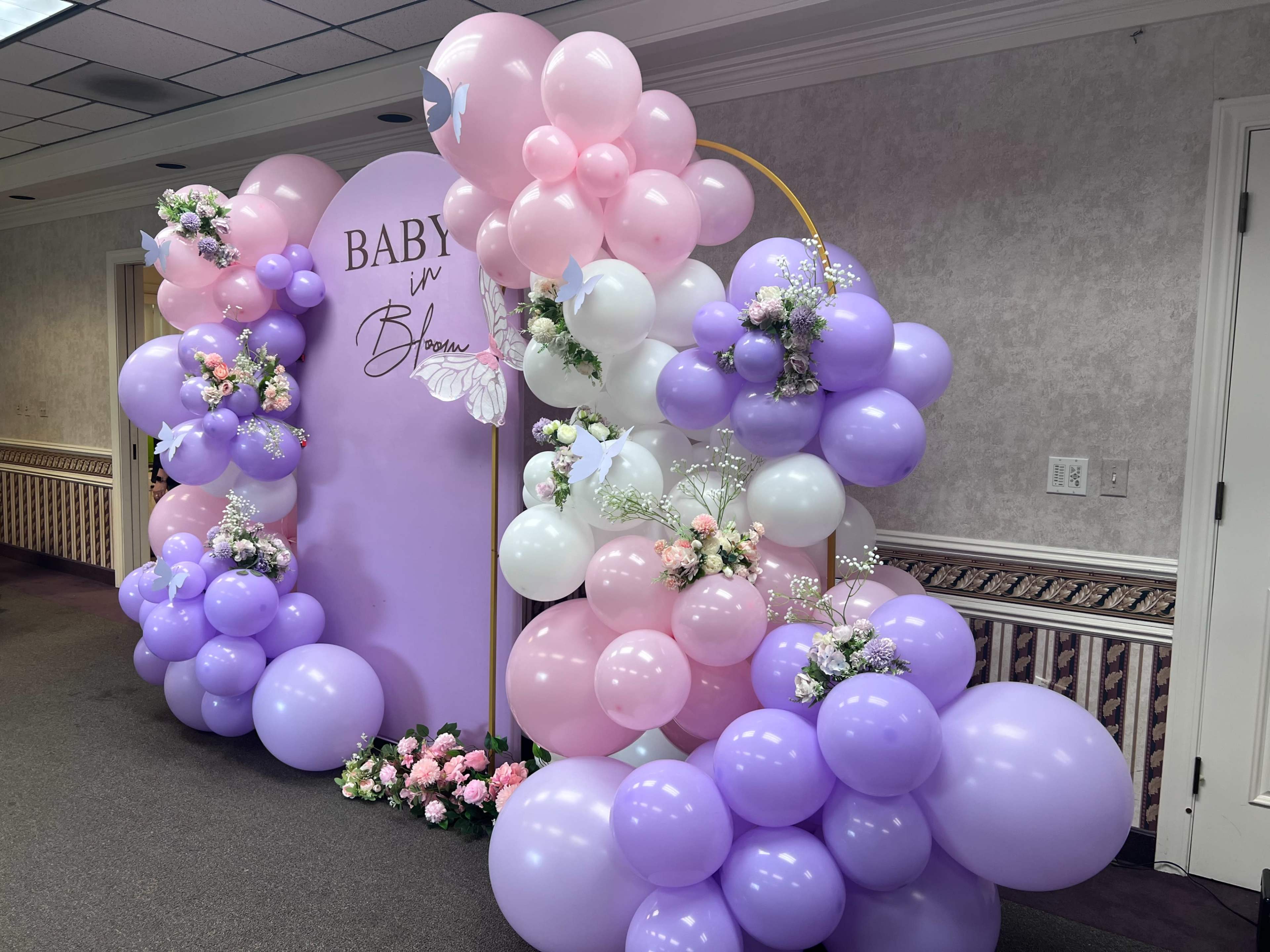 A decorative balloon arch with pink, purple, and white balloons, arranged around a backdrop that reads "BABY in Bloom," with floral accents.
