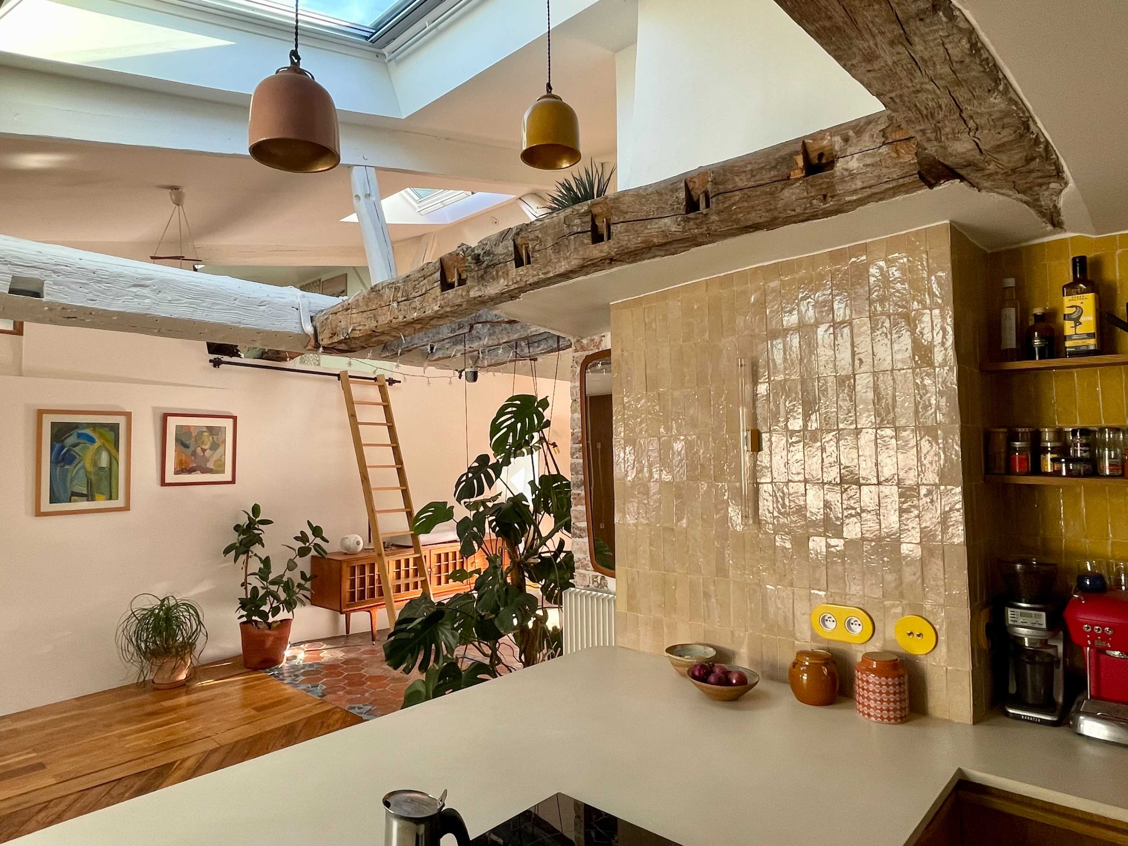 The image shows a bright, open kitchen area with wooden beams, a modern countertop, and plants, alongside a living space featuring framed artwork and a ladder leading to an upper level.