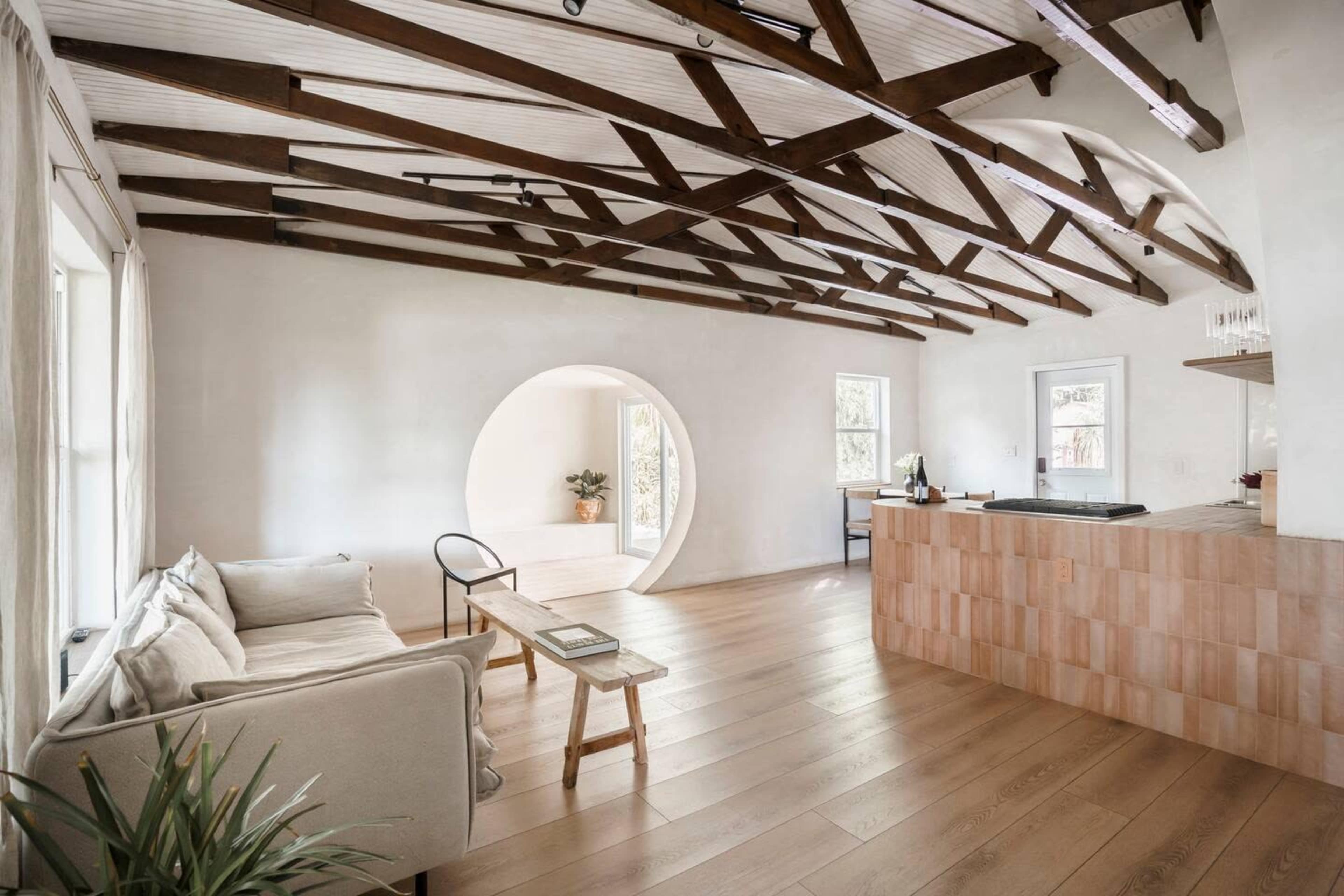 The image shows an interior living space featuring exposed wooden beams, a light-colored sofa, a small table, and a kitchen area with a tiled counter.