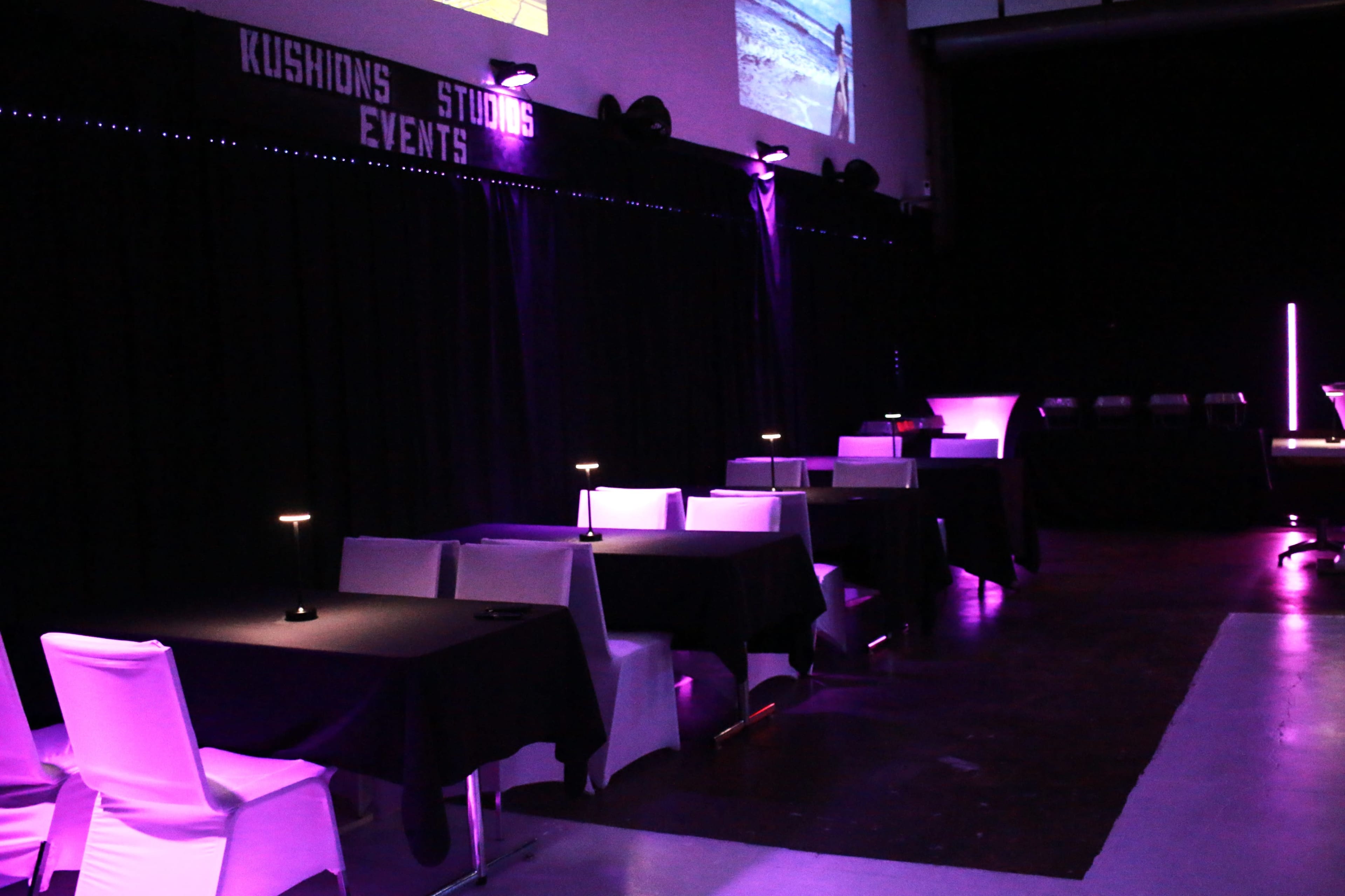 The image shows a dimly lit event space featuring black tables with white chairs, illuminated by purple lighting, along with a black backdrop displaying the words "Kushions Studios Events."