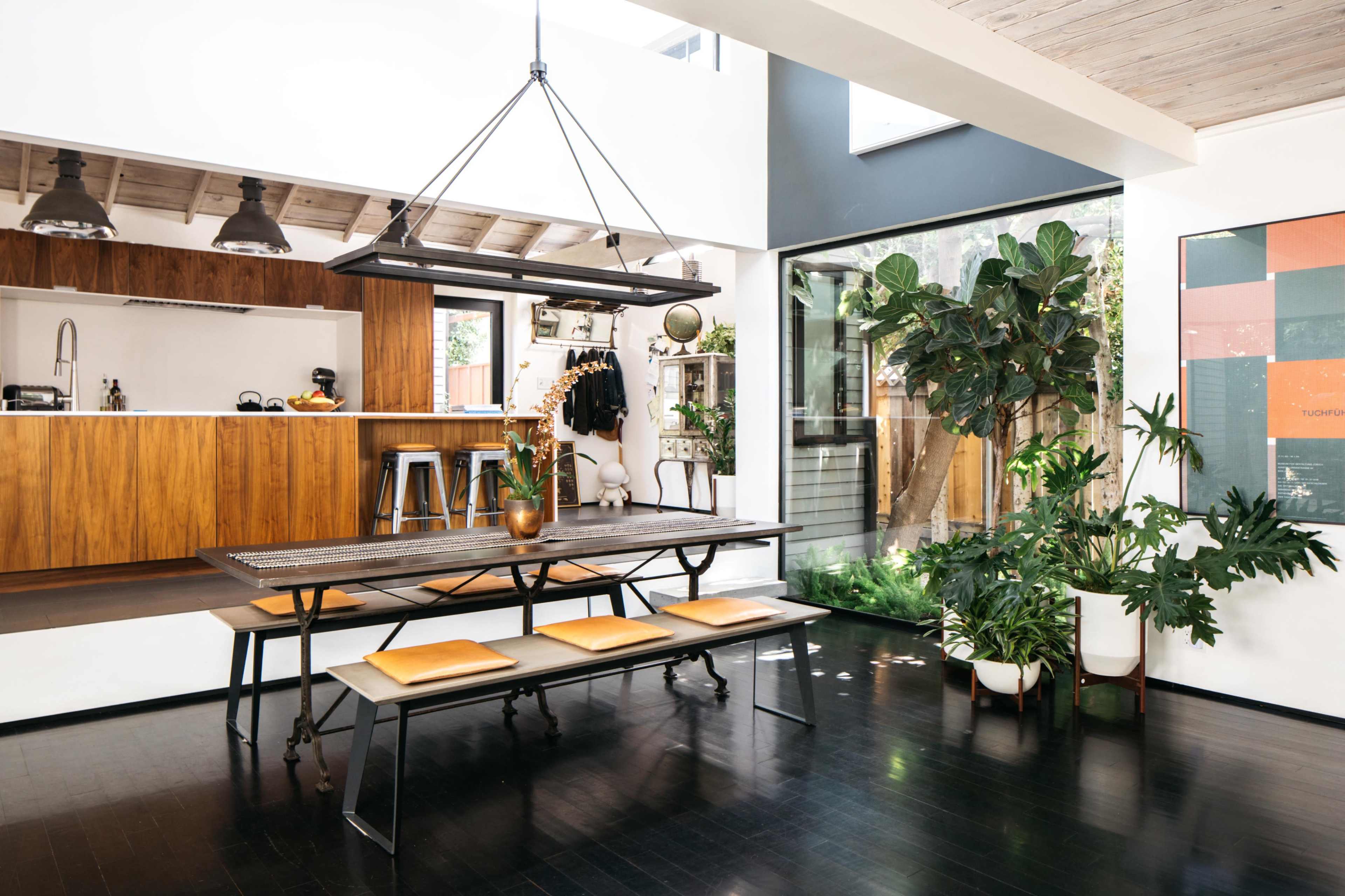 A modern dining area with a rectangular metal table, wooden benches, and plants, adjacent to a kitchen with wooden cabinetry and a large window.