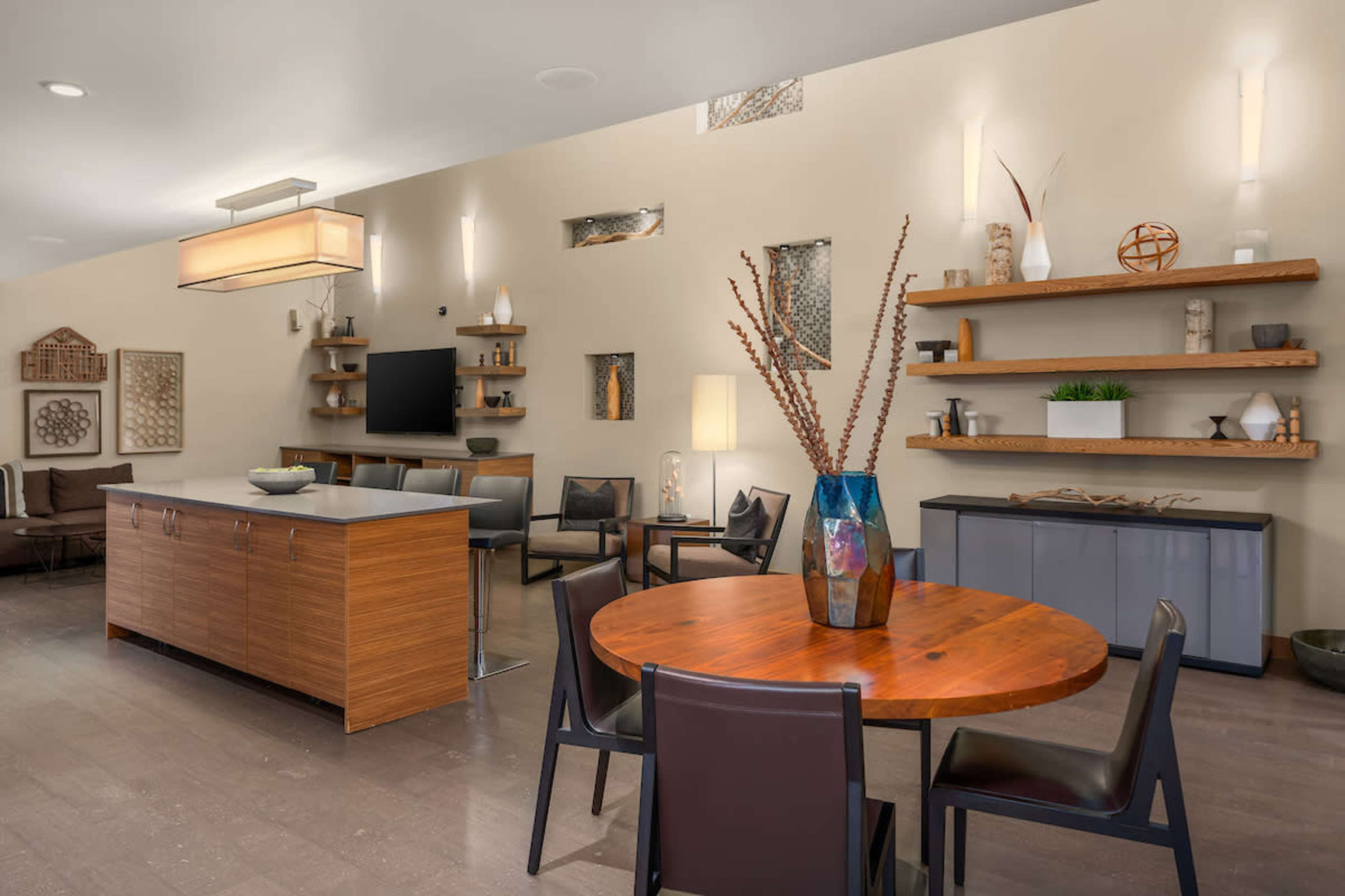 A modern living space features a wooden dining table with chairs, a kitchen island, and shelves displaying decorative items against a neutral wall.