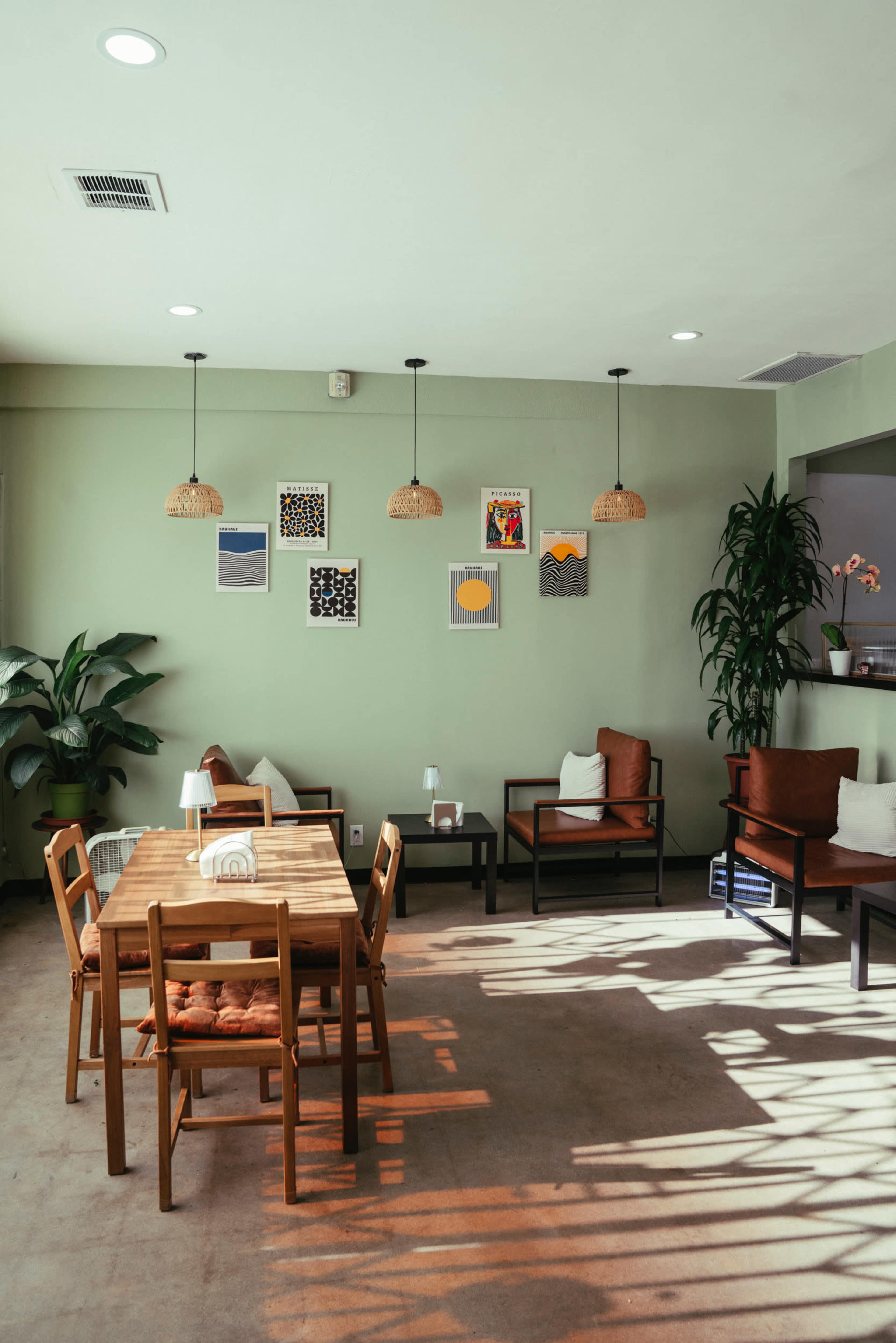 A cozy indoor space features a wooden table with chairs, a few armchairs, and artwork on the green walls, illuminated by pendant lights.