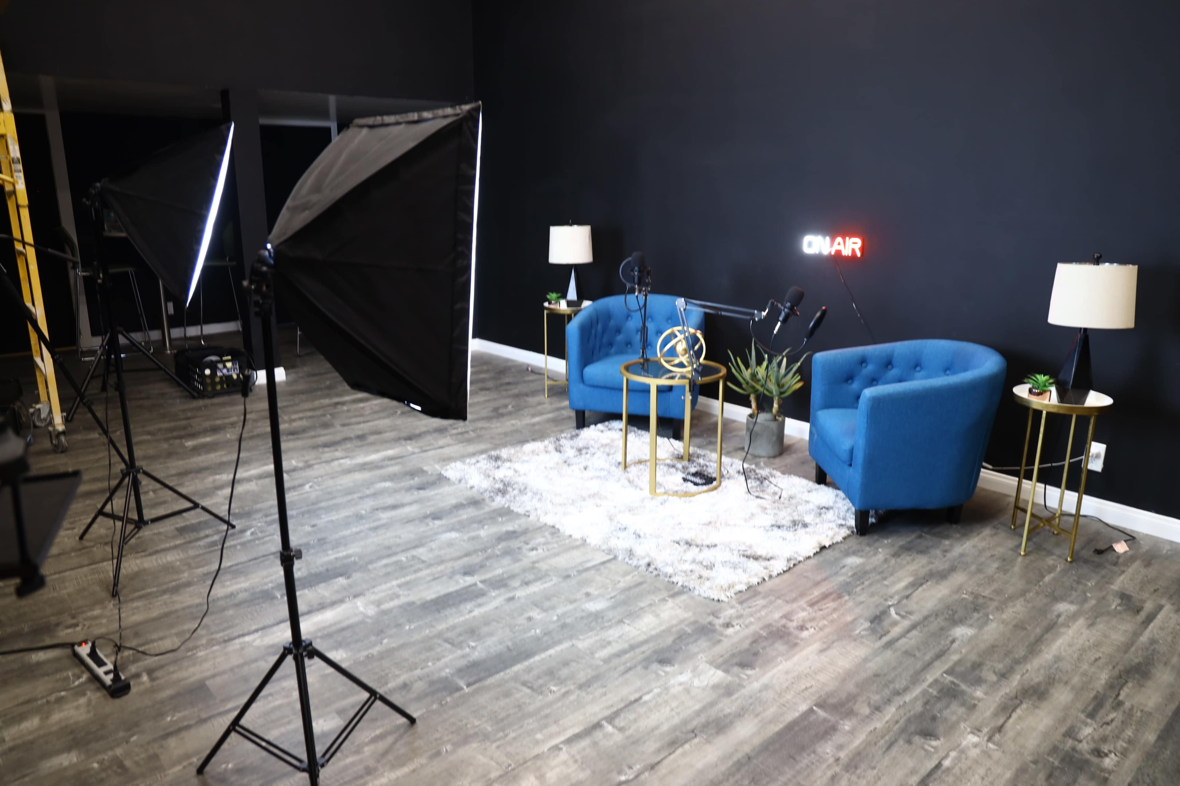 The image shows a small recording studio featuring two blue chairs, a table, a rug, a plant, and lighting equipment, all against a black wall with an "ON AIR" sign.