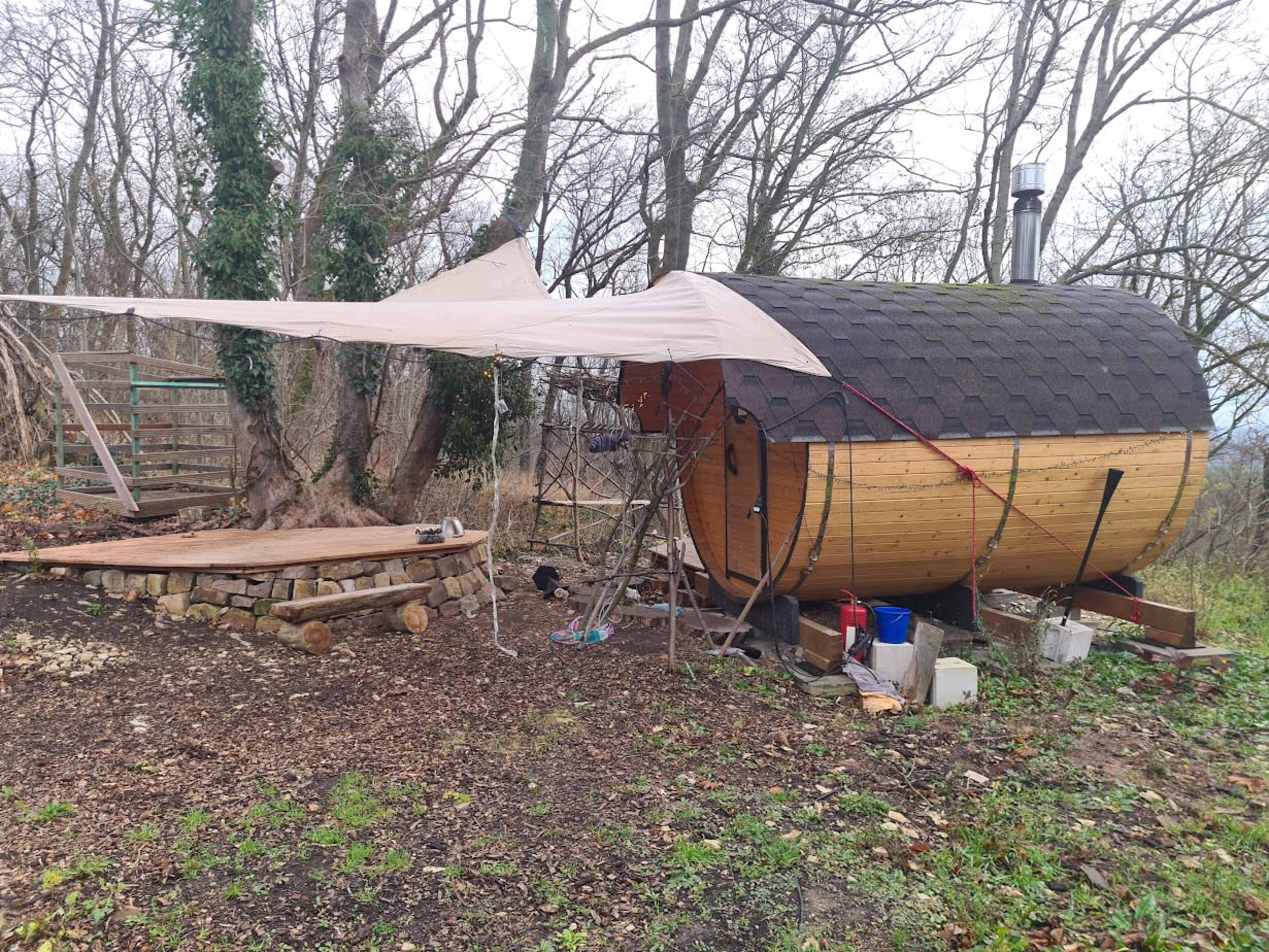 The image shows a round wooden structure resembling a sauna, with a tent-like cover extending beside it, set in a wooded area with leafless trees.